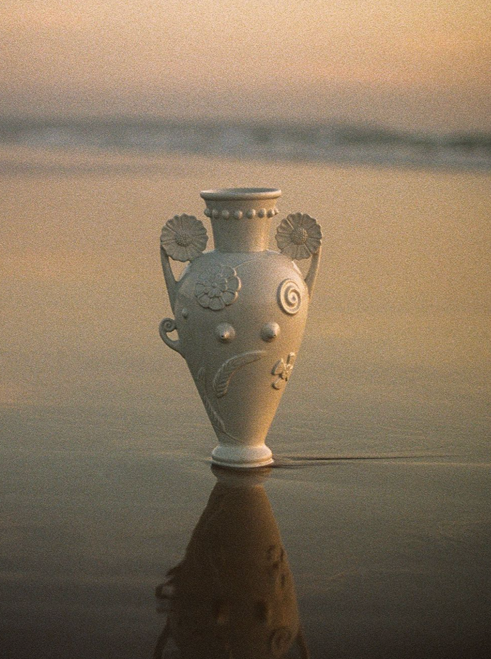 A decorative White Earthenware Vase by Lobjet, with floral patterns reminiscent of a Greek Amphora, stands on a reflective wet sand surface at the beach. The background features a soft-focus view of the ocean and a pastel-colored sky at either sunrise or sunset.