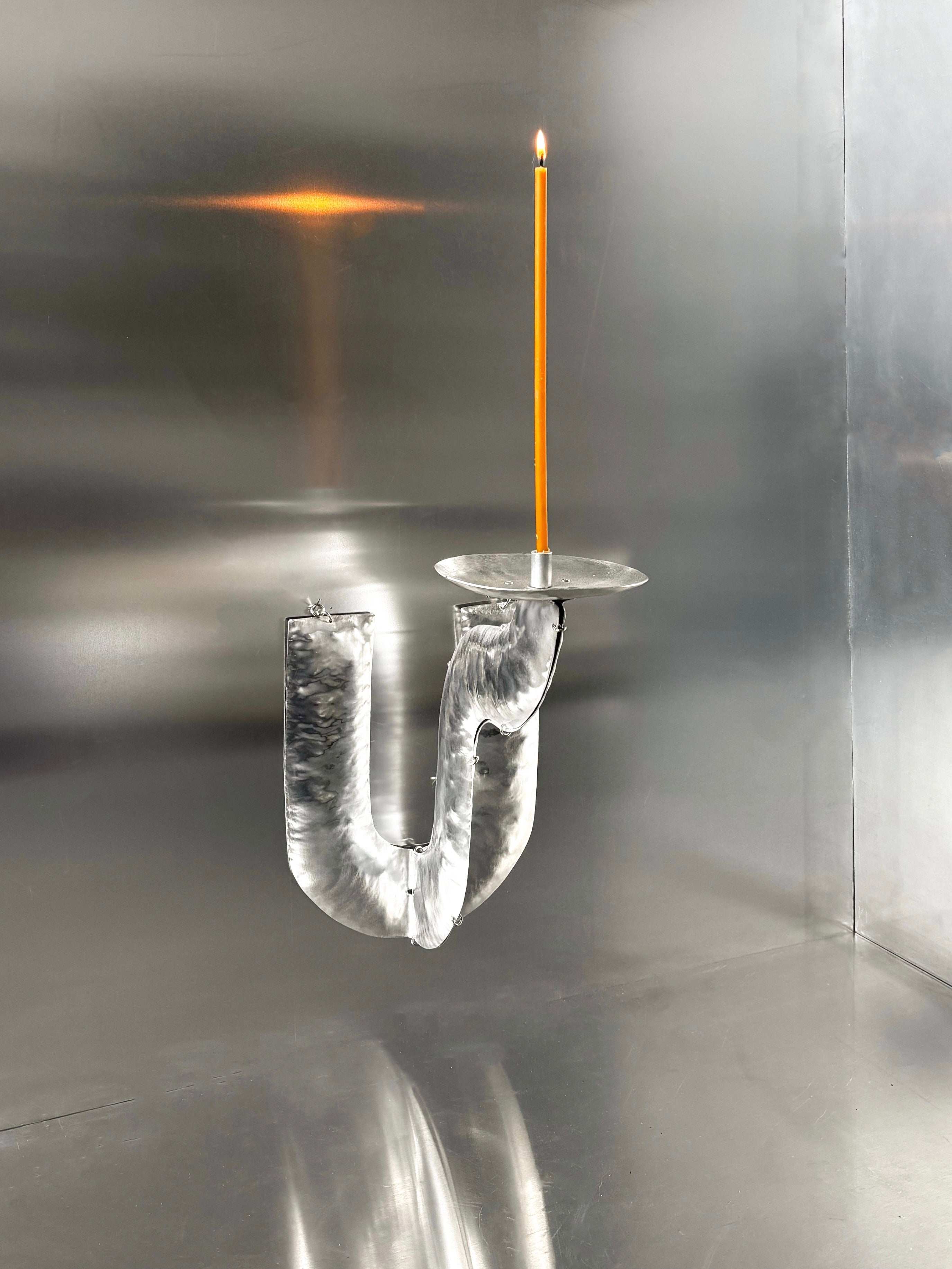 The Phaedra Candle Holder Wall by Bregje Sliepenbeek features a lit orange candle on an upright, reflective U-shaped aluminum holder against a shiny silver backdrop, creating a minimalistic and modern look.