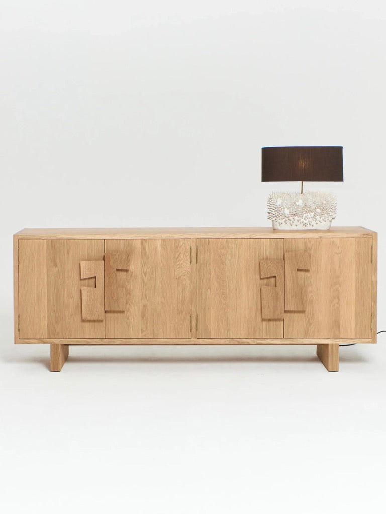 The "Douro" Cabinet in Oak by Project 213A, a wooden sideboard with geometric handles, stands on short legs. Crafted from solid oak, it boasts hand-carved details. Atop the cabinet sits a decorative black table lamp with a detailed coral-like base. The background is plain white.