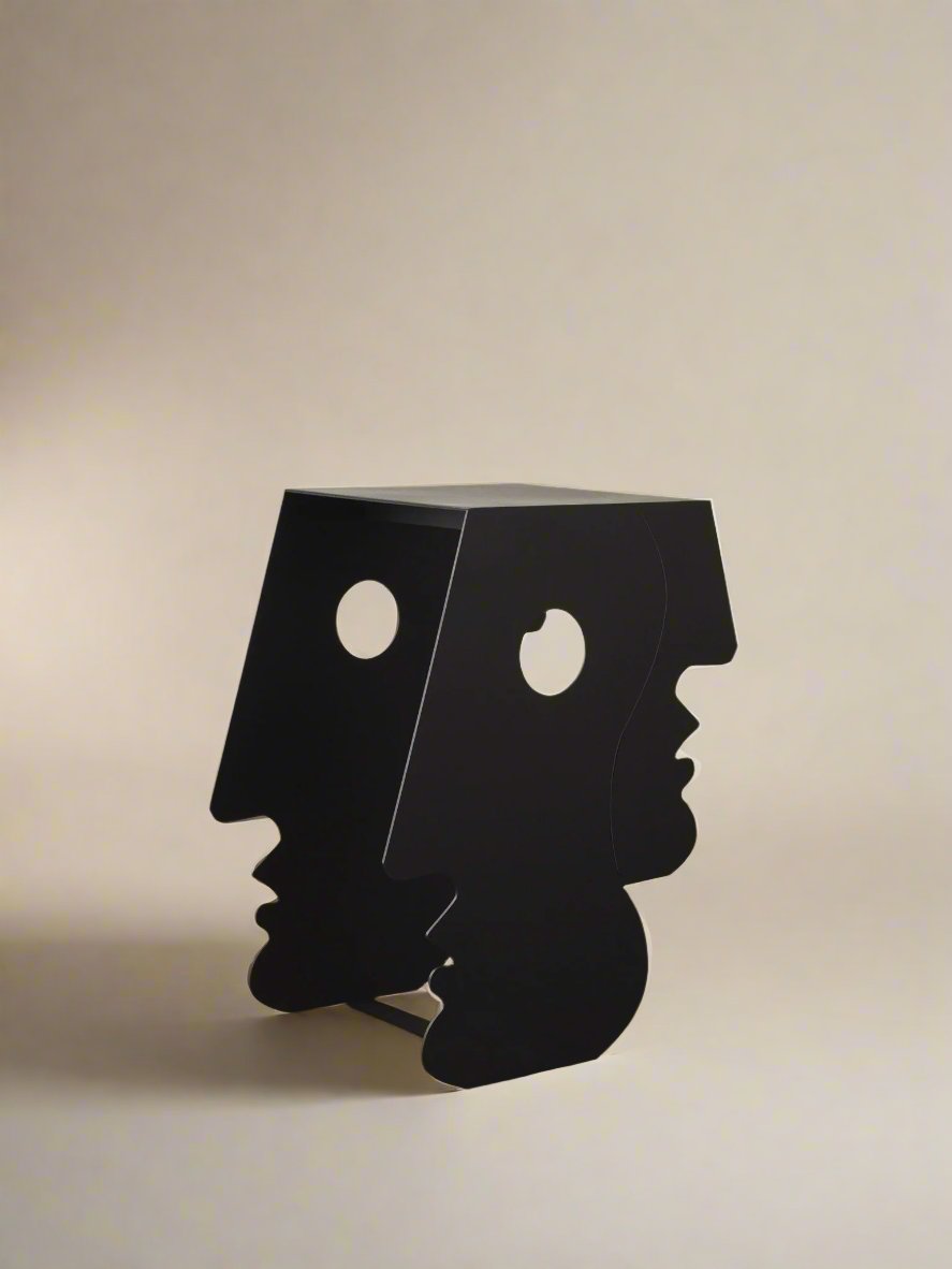 The "Lucia" Side Table in Black by Animate Objects features a modern, artistic design crafted from laser-cut iron. Its profile faces on either side and circular cutouts for eyes create a distinctive look, while it stands with elegance on a polished brown floor against a simple backdrop, evoking the style of Bauhaus photography.