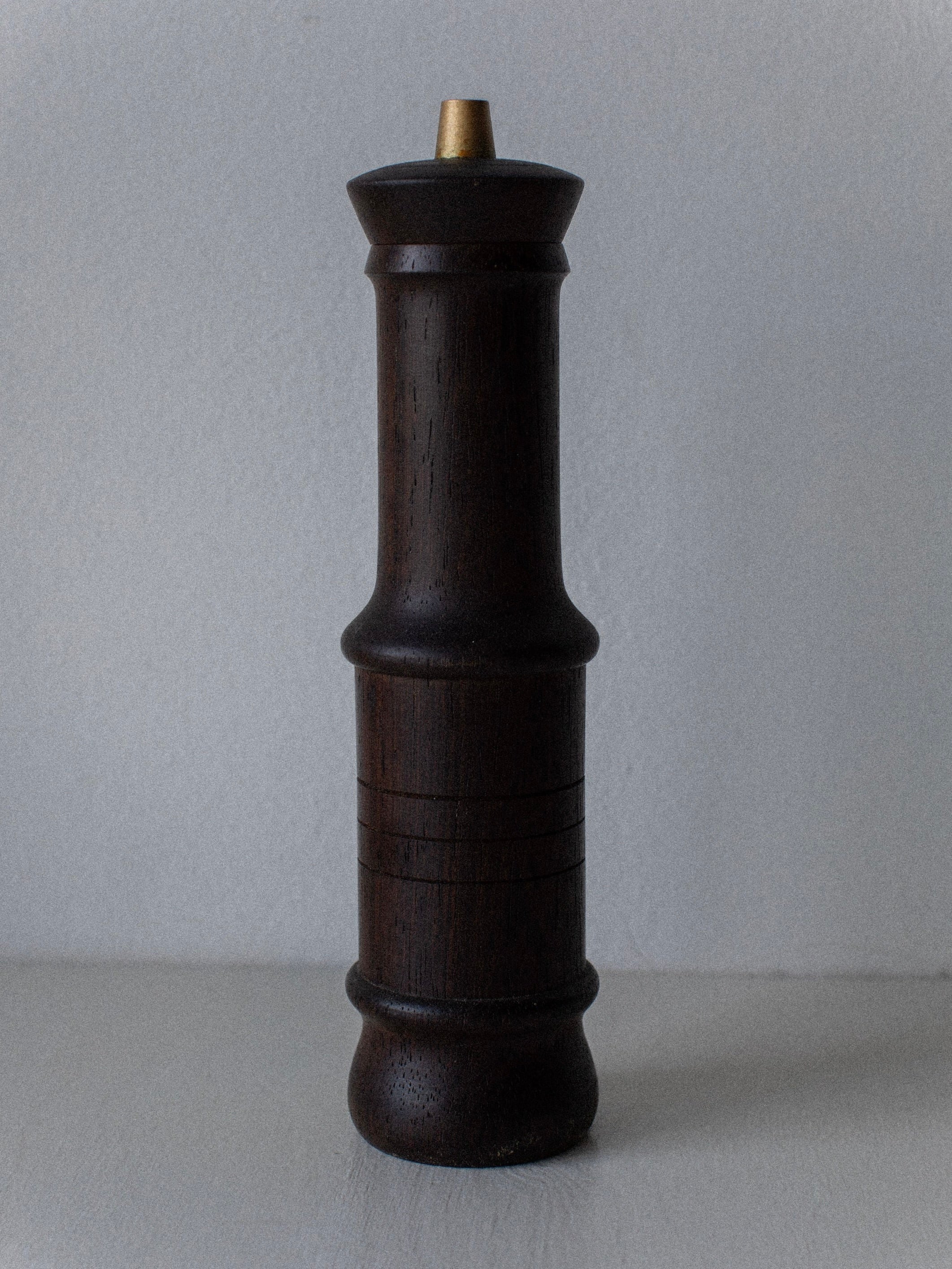 The Wenge Wood Grinder by Out For Lunch stands tall against a light gray background, showcasing its dark brown wenge wood body and golden top knob. The pepper mill, with smooth, slender, and slightly rounded contours, features engraved horizontal lines in the middle section. A vintage grinder of Danish design.