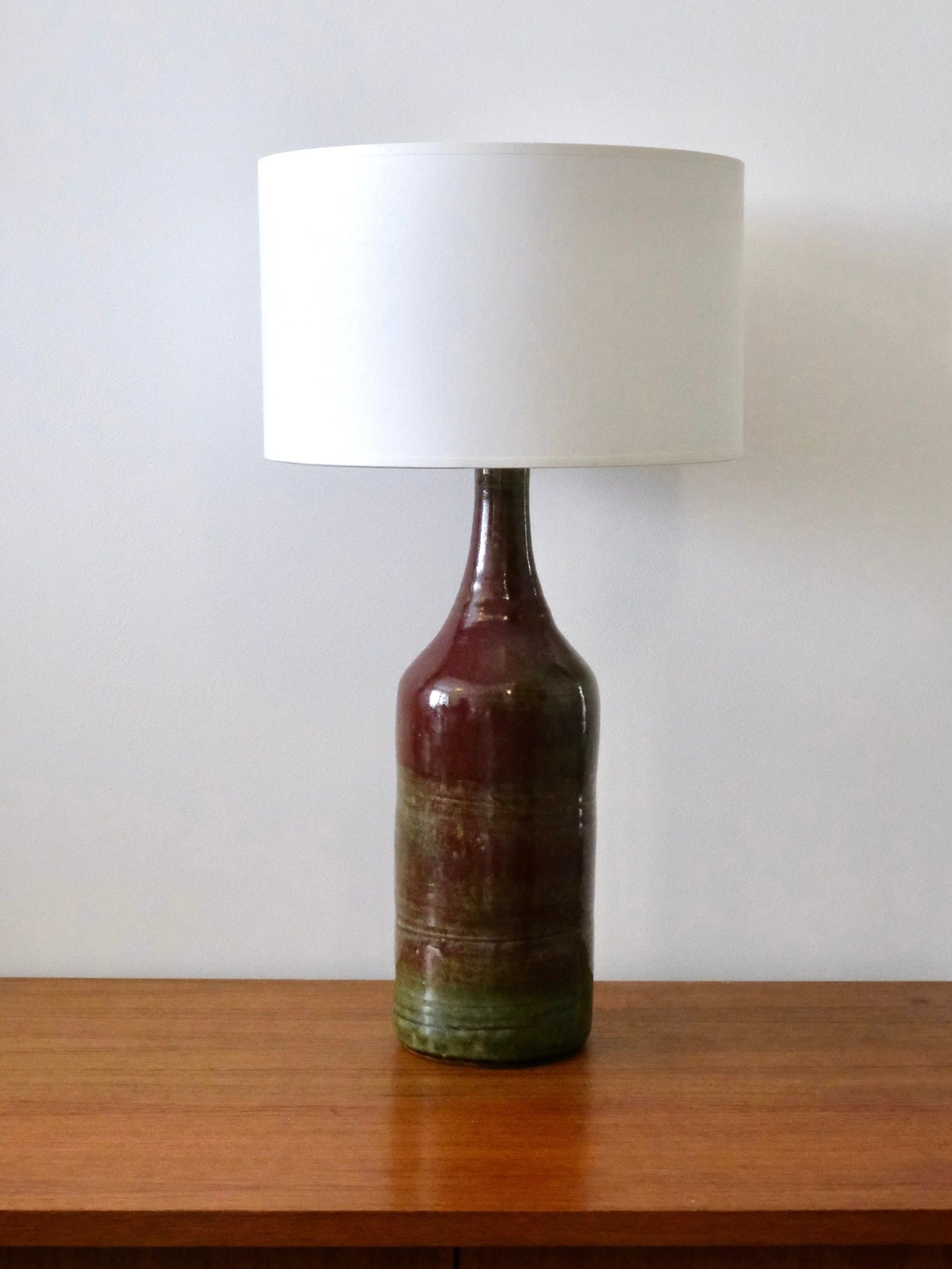 A 70s-inspired Ceramic Lamp by Fongombault from LA NOUVELLE GALERIE features a bottle-shaped base in earthy brown and green tones on a wooden surface, topped with a white cotton lampshade. Its design reflects the simplicity of Abbaye de Notre-Dame de Fongombault against a plain white background.