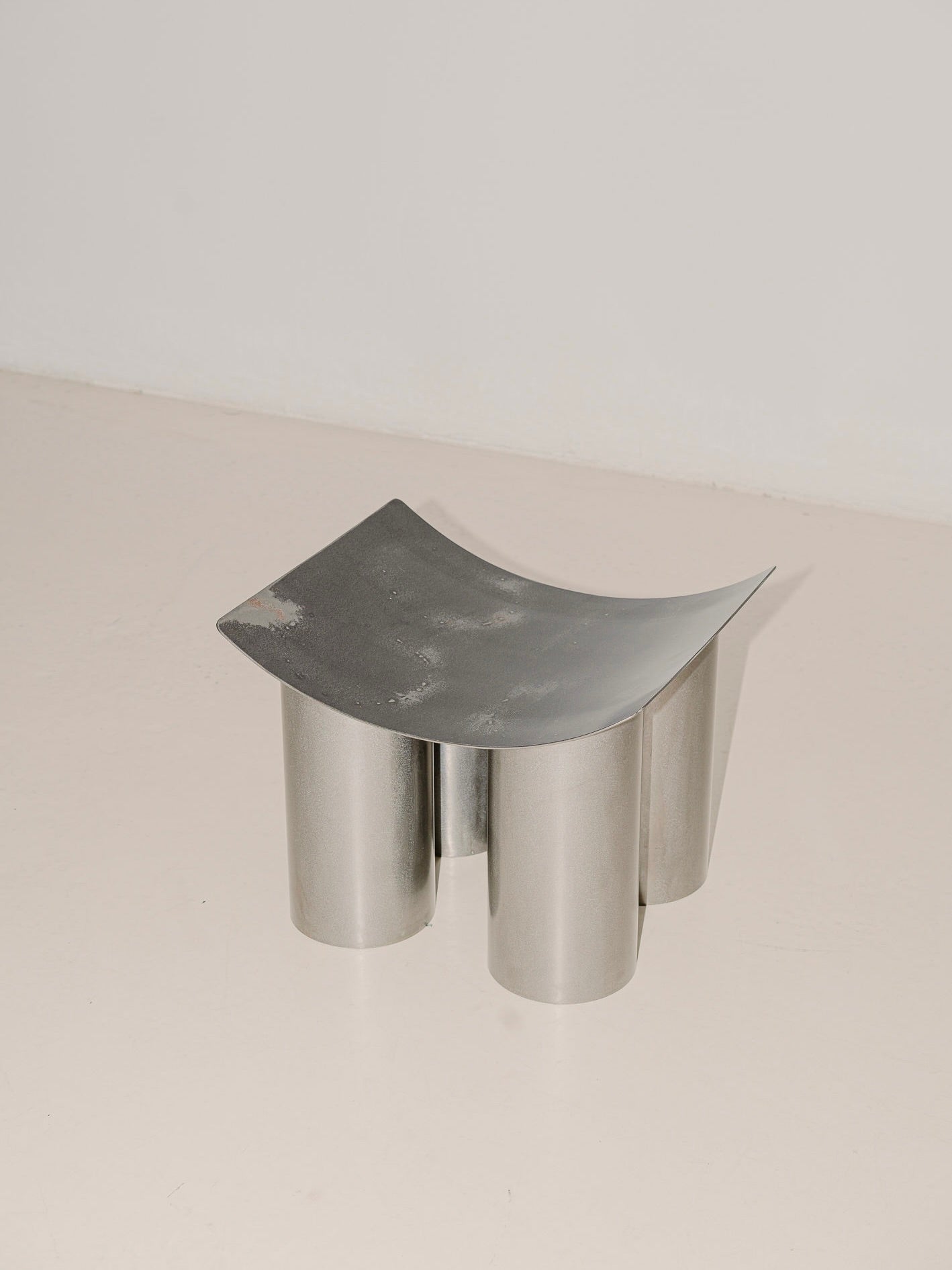 The Good Living&Co.'s "Tut" Stool in Stainless Steel boasts a modern, minimalist design with a curved seat and four cylindrical legs. It stands out beautifully against a plain, light-colored background, highlighting its contemporary charm.