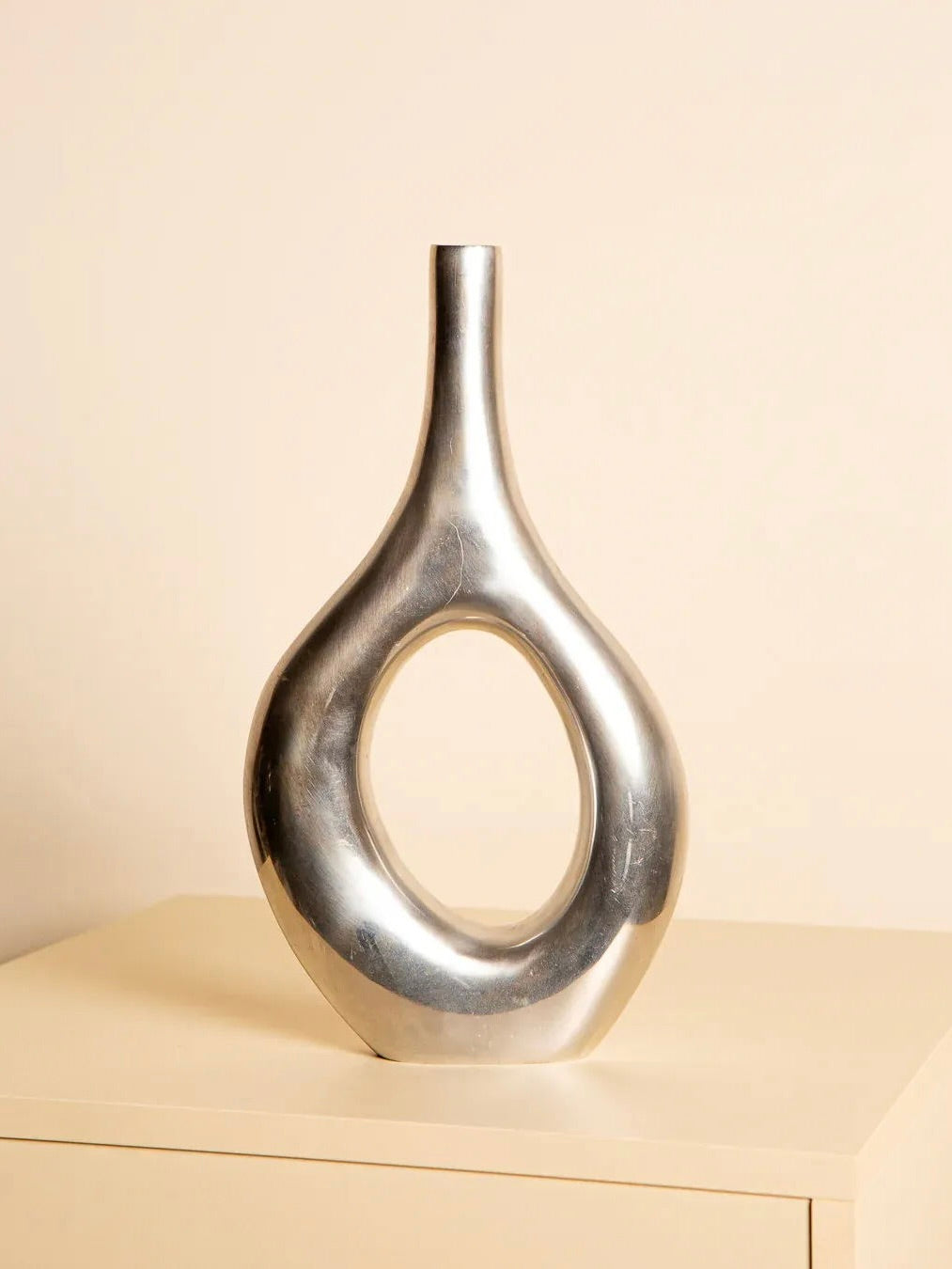 The Long Italian Vase in Cast Aluminum 80's by Treaptyque stands on a light surface against a beige background. With an open-centered, smooth teardrop shape, the metallic vase reflects the modern artistry of Northern Italy's Como region, showcasing contemporary elegance.