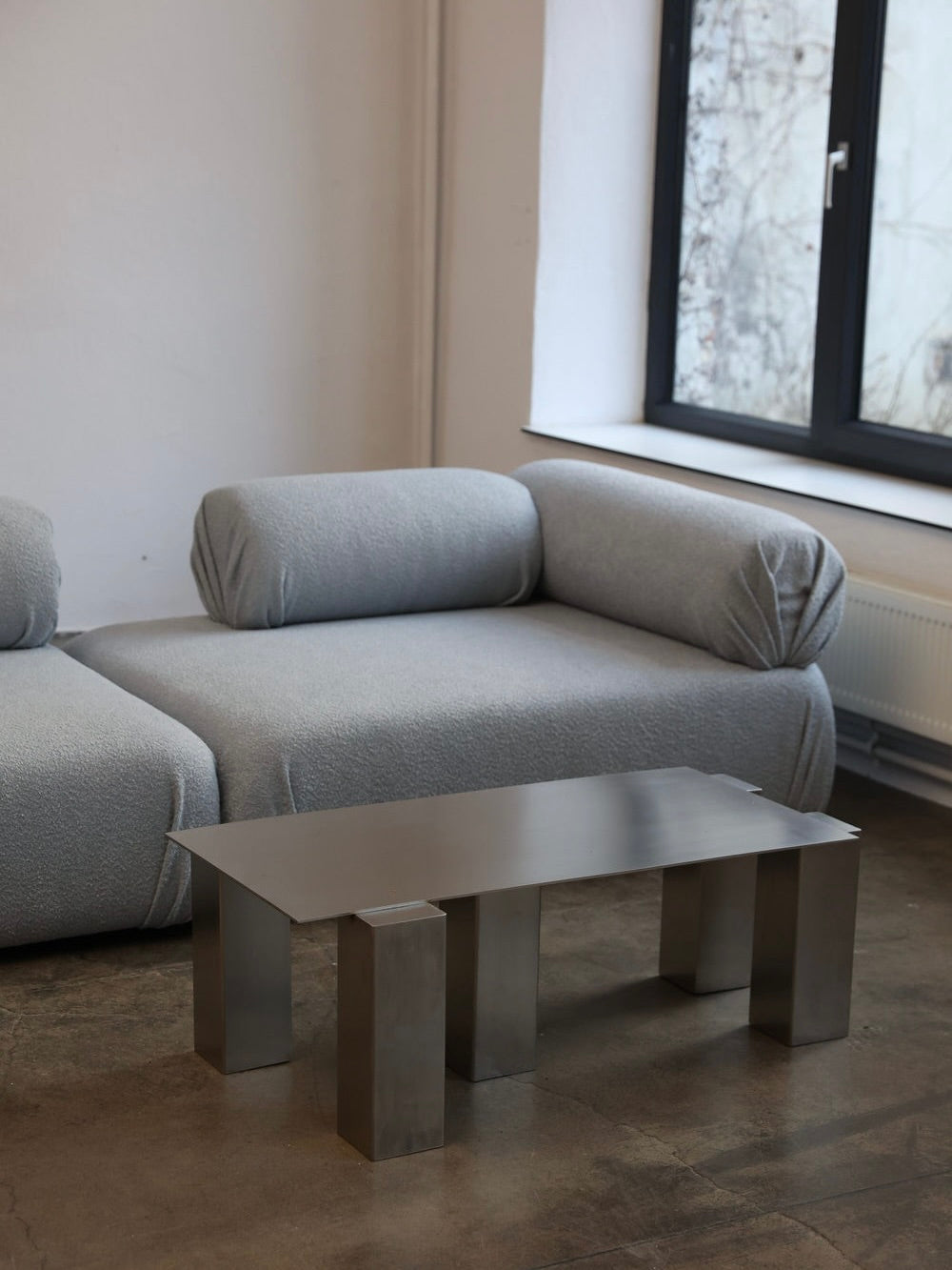 A modern minimalist living room features a sleek gray sofa with cylindrical cushions facing The Good Living&Co.'s Atrium Coffee Table, distinguished by its unique block legs. A large window with black trim provides a view outside, while concrete flooring and stainless steel accents enhance the industrial vibe.