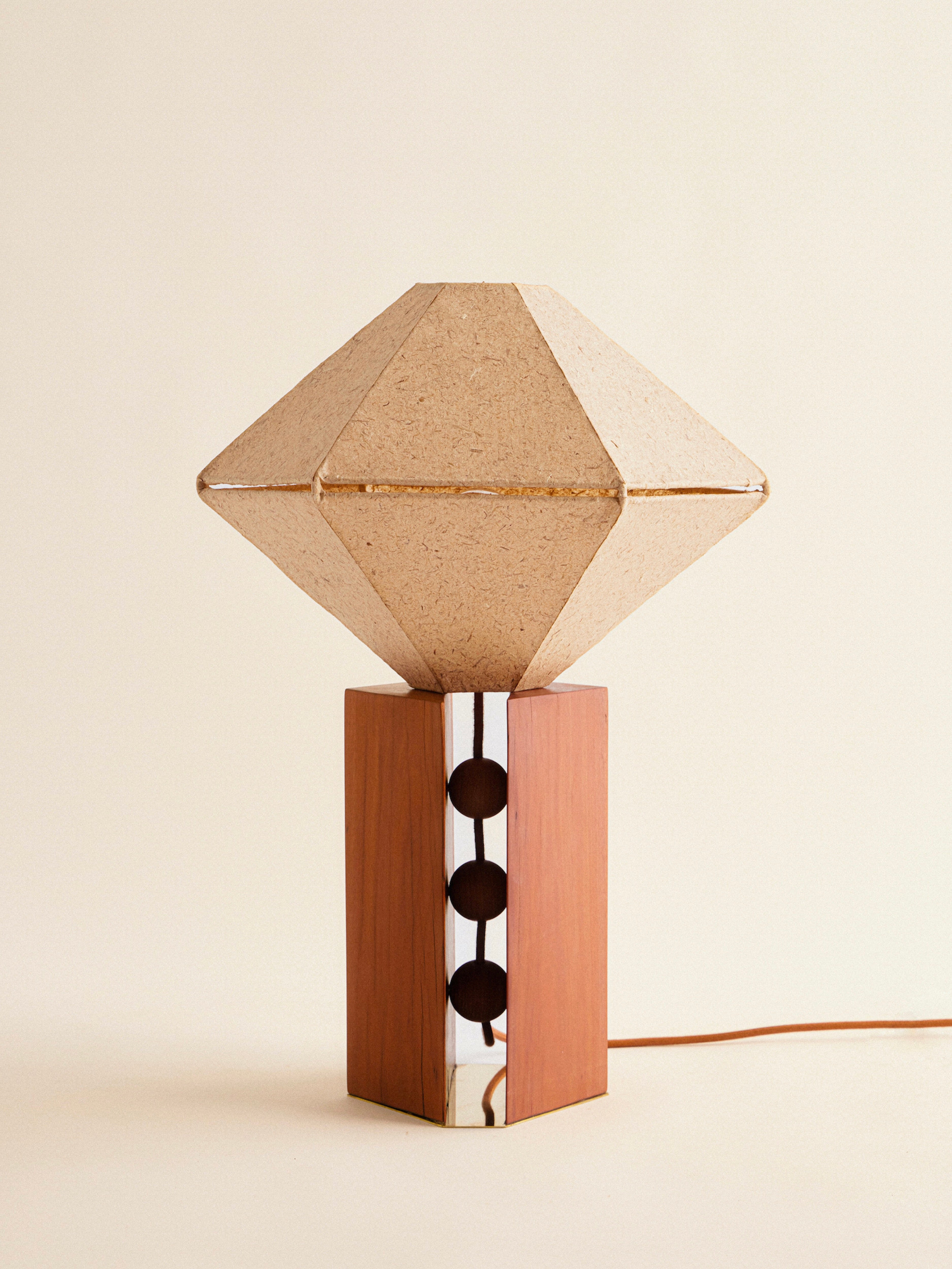 Discover the "USO" Table Lamp by Lana Launay, a handcrafted piece from Australia that captures the essence of Googie architecture. It features a geometric silhouette and a textured beige shade reminiscent of a cut gem, while resting on a wooden base with circular cutouts and metallic accents. An orange power cord adds the perfect finishing touch against its neutral backdrop.