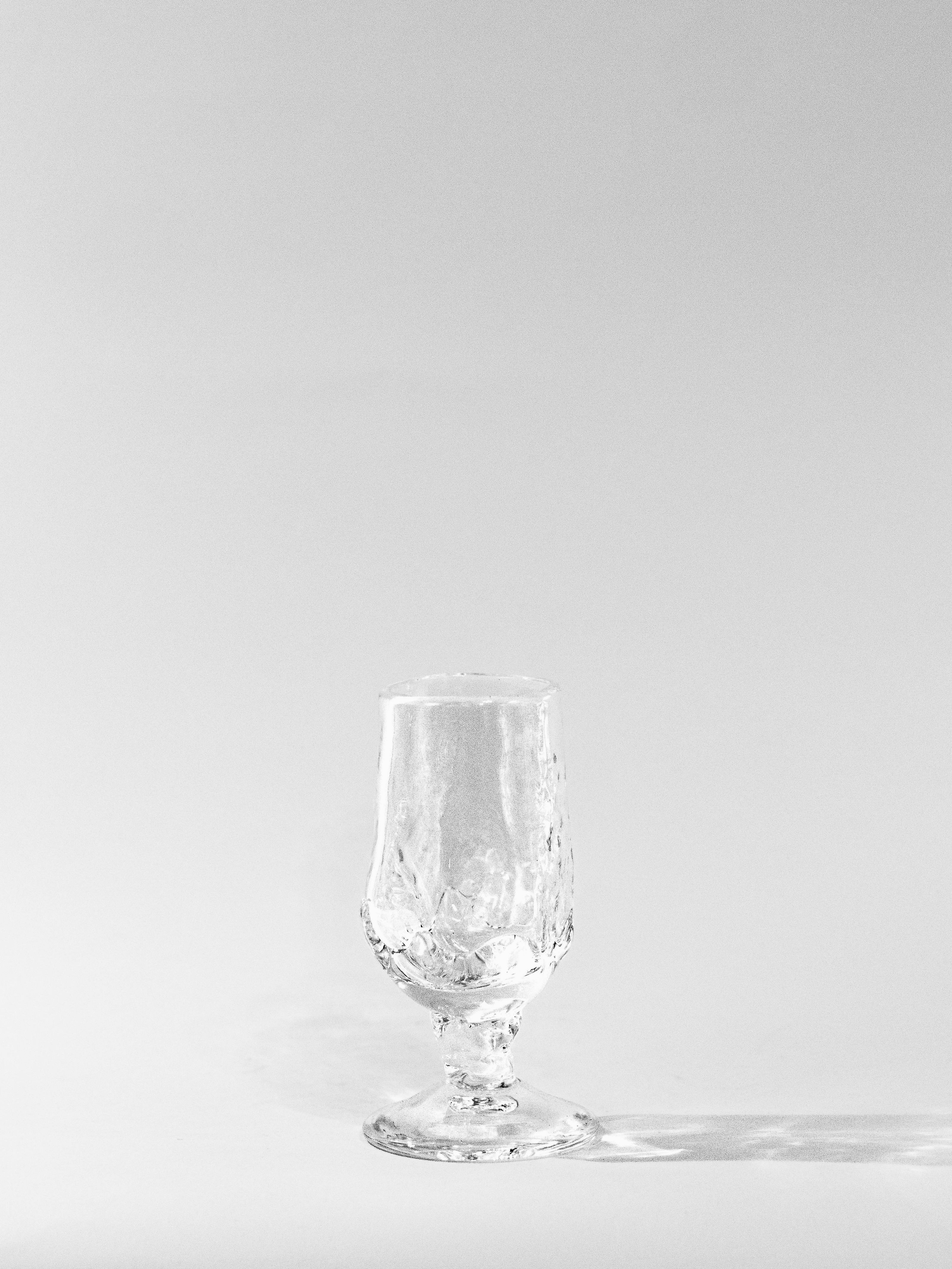 A Footed Goblet from SzkĆo Studio, made of clear, lead-free soda-lime glass, stands on a plain white background. The freehand-blown design features a textured surface where lighting creates soft reflections and shadows, highlighting its intricate details.