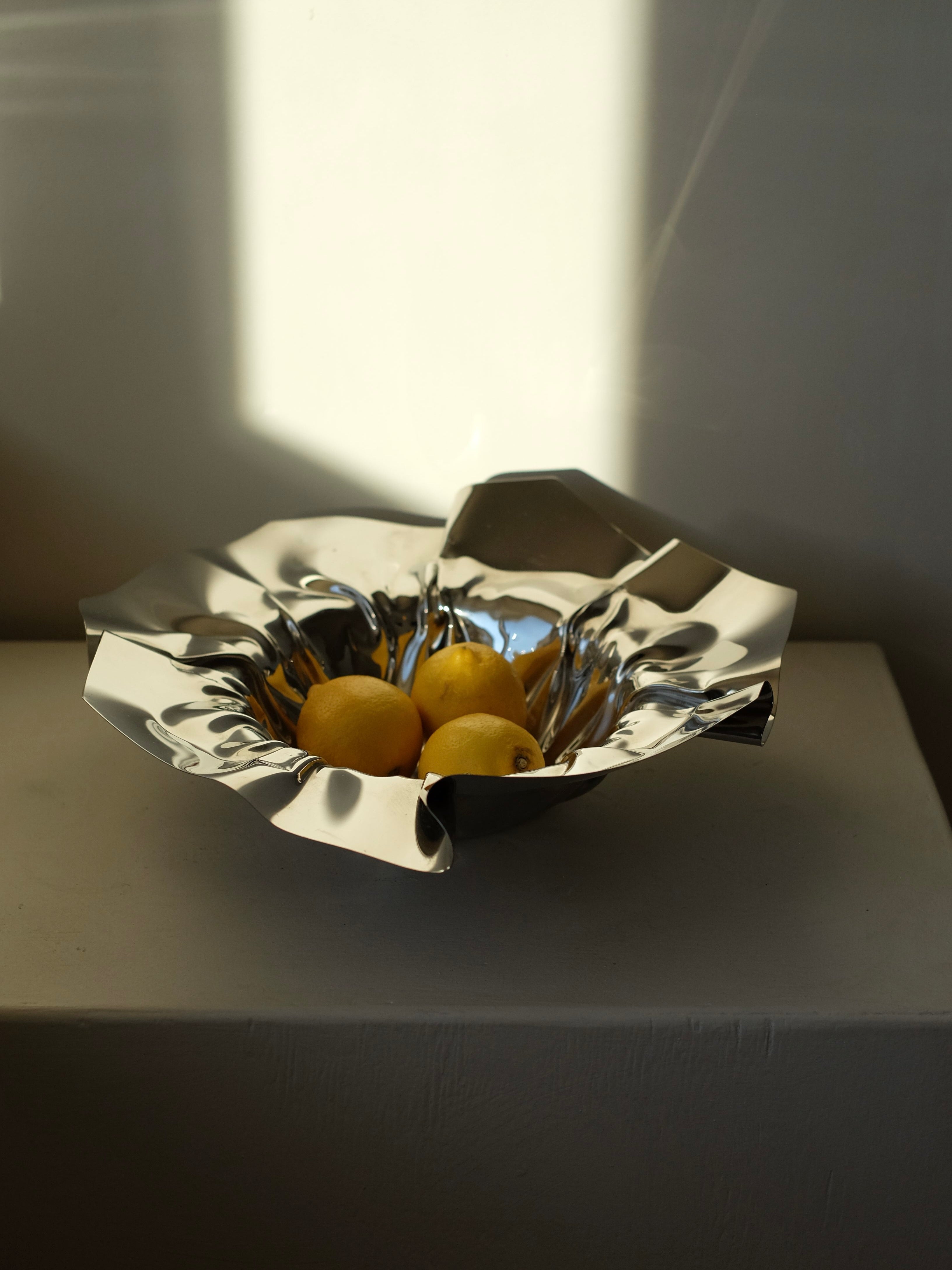 The Folded Chrome Bowl by septembre studios, with its shiny metallic finish and unique folded shape, elegantly holds three lemons. Placed on a white surface, the bowl is beautifully illuminated by soft natural light, creating delicate shadows on the wall behind—making it a perfect accent for any interior space.