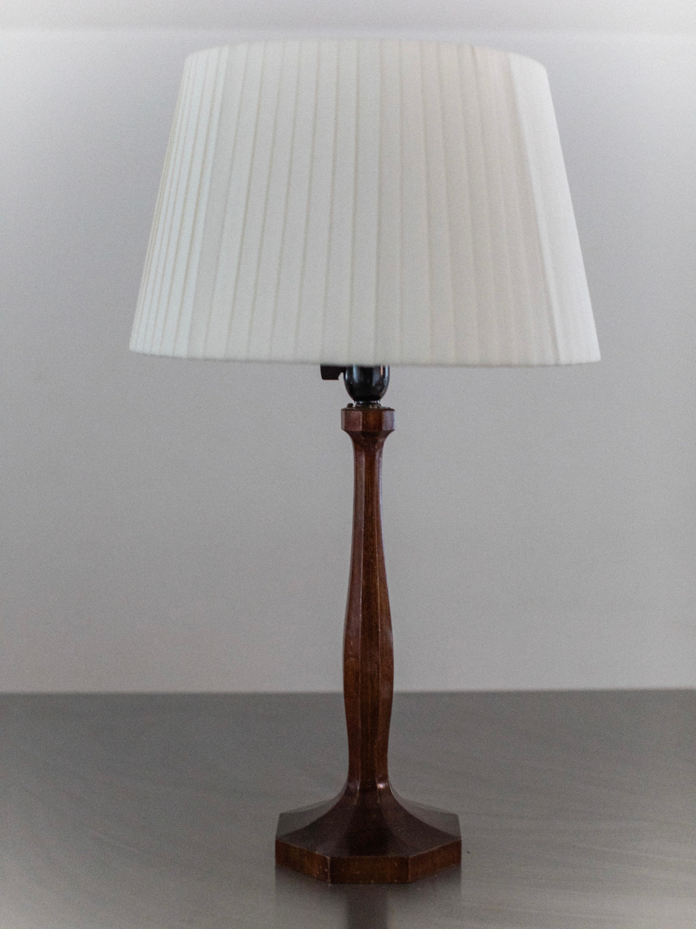 The 1950's Table Lamp by Out For Lunch boasts a minimalist design with a tall, dark wooden base and a white pleated lampshade, elegantly set on a sleek surface against a simple backdrop.