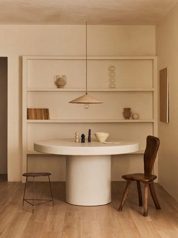 A minimalist dining area features the "Persillé" Dining Table by MarlotBaus with mismatched chairs. A simple pendant light highlights the table's handmade technique, while a built-in shelf displays neutral-toned decor, books, and ceramics against light wood flooring.