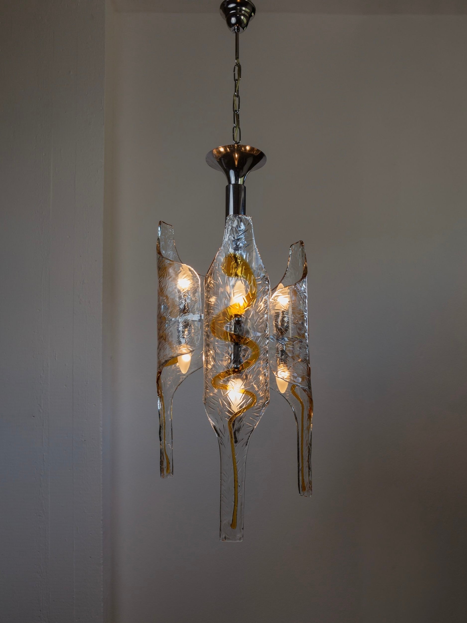 The Vintage Murano Glass Chandelier by Toni Zuccheri for Mazzega, offered by Bottega Jacobs, features a sleek metallic structure adorned with wavy, textured Murano glass panels. Its design provides a warm glow from the ceiling.