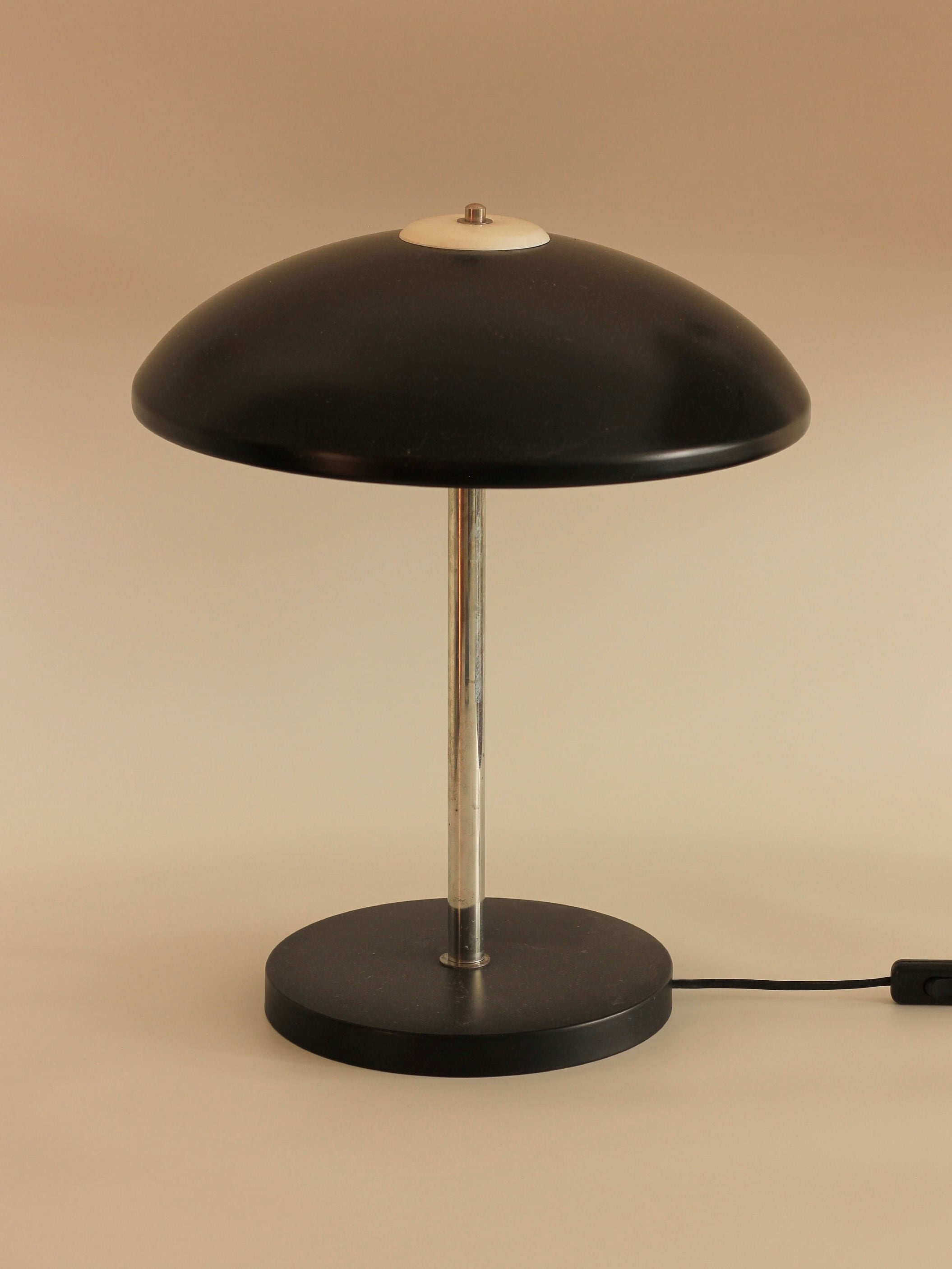 Introducing the Mushroom Table Lamp from Boga Avante Shop, featuring a modern black design with a round base and a smooth, conical shade. The lamp boasts a sleek chrome stem and exhibits minimalistic aesthetics, with the power cord visibly extending from the base to the right. Perfect for mid-century modern interiors, it is showcased against a plain and neutral background.
