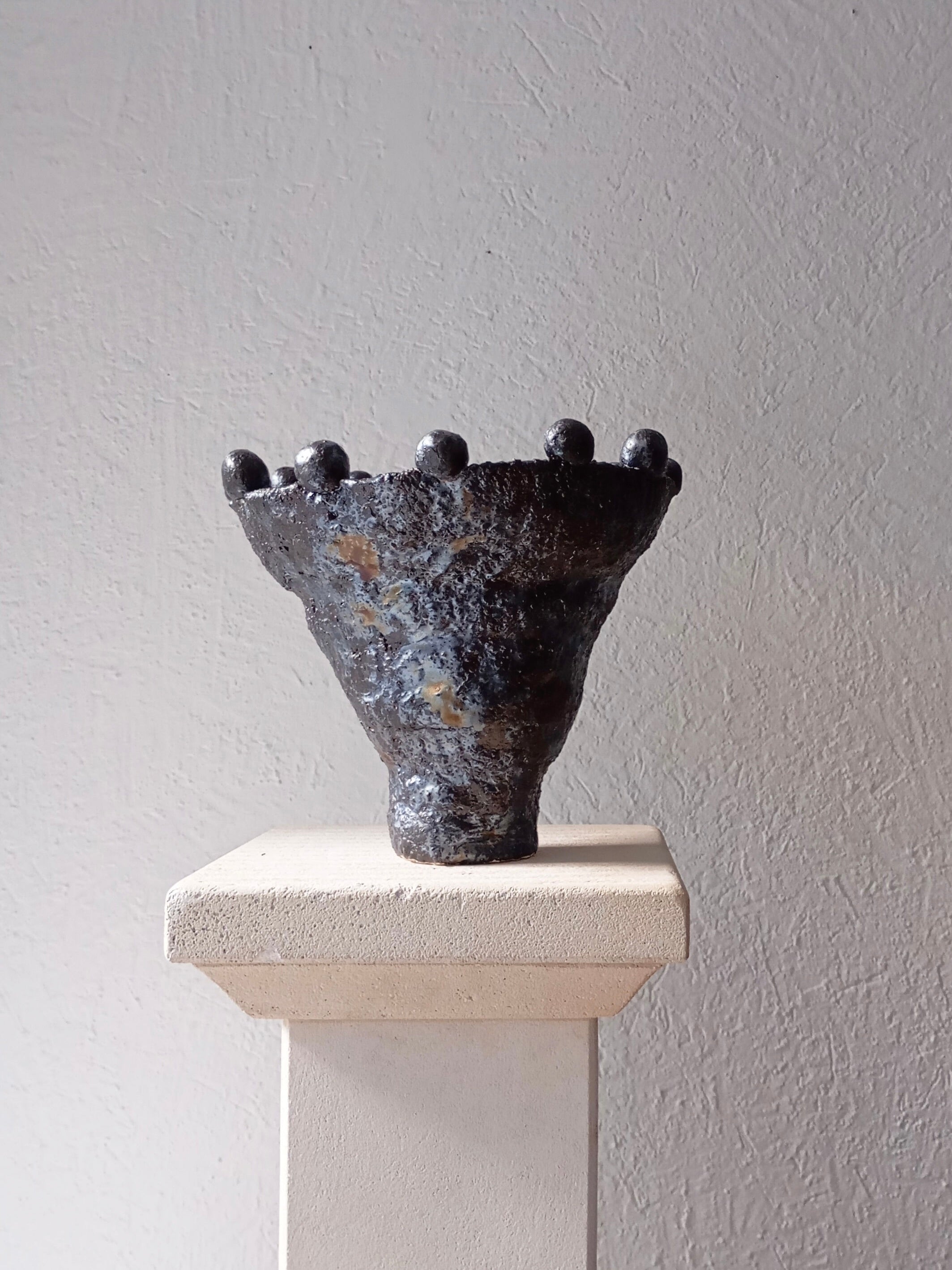 The "Coupe Lilibeth 2" by Claire Pain is a textured dark ceramic vase with a wide rim and small rounded protrusions, featuring a subtle metallic glaze. It elegantly sits on a light-colored pedestal against a plain textured wall, making it perfect for decorative use.