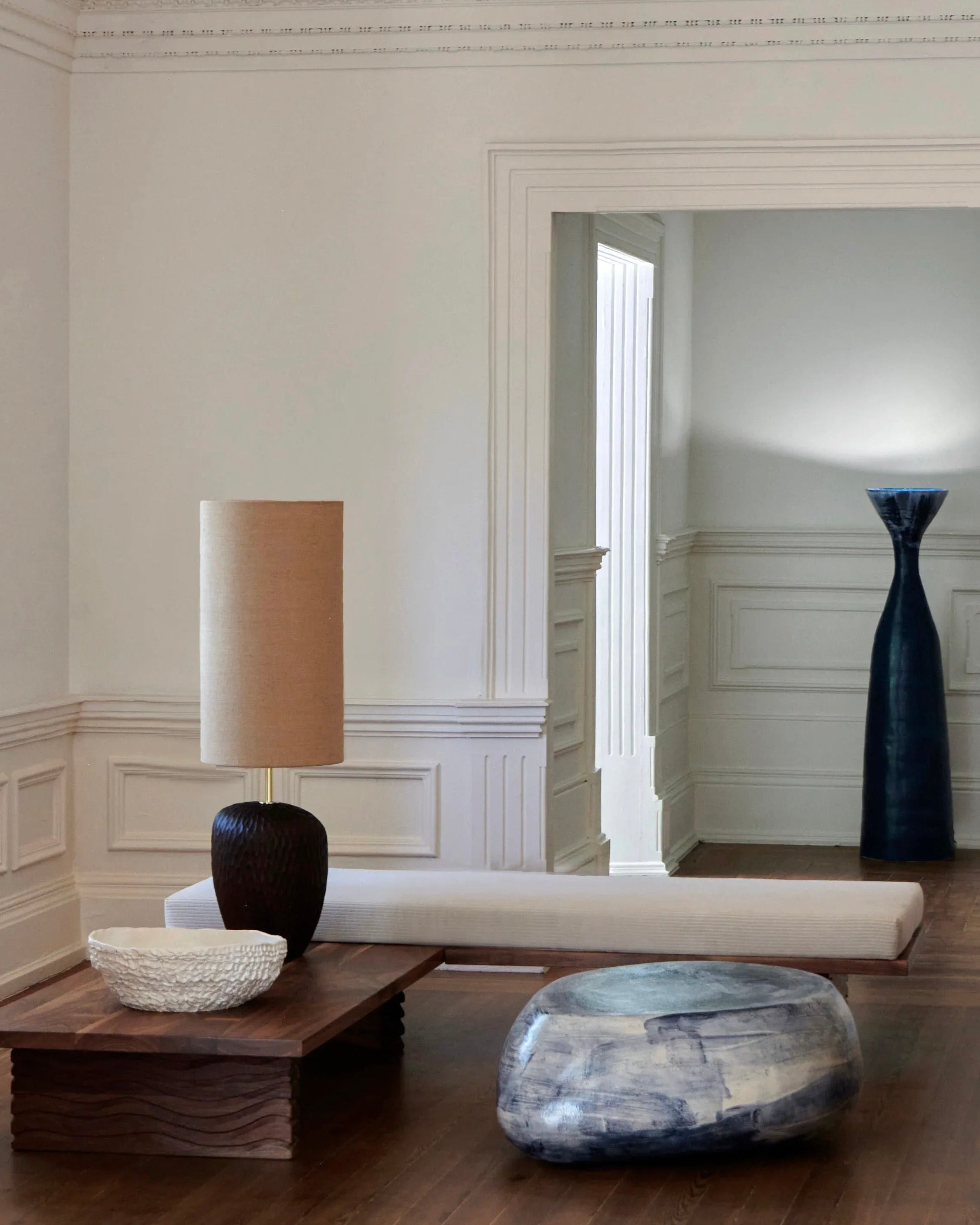 A minimalist room with white walls features a low wooden coffee table adorned with a beige lamp and a decorative bowl. The Nazaré Daybed L Shape by Project 213A, known for its modern design and comfort, sits nearby. A large, blue vase is visible in the adjacent hallway through the rectangular doorway, adding a touch of color to the serene space.