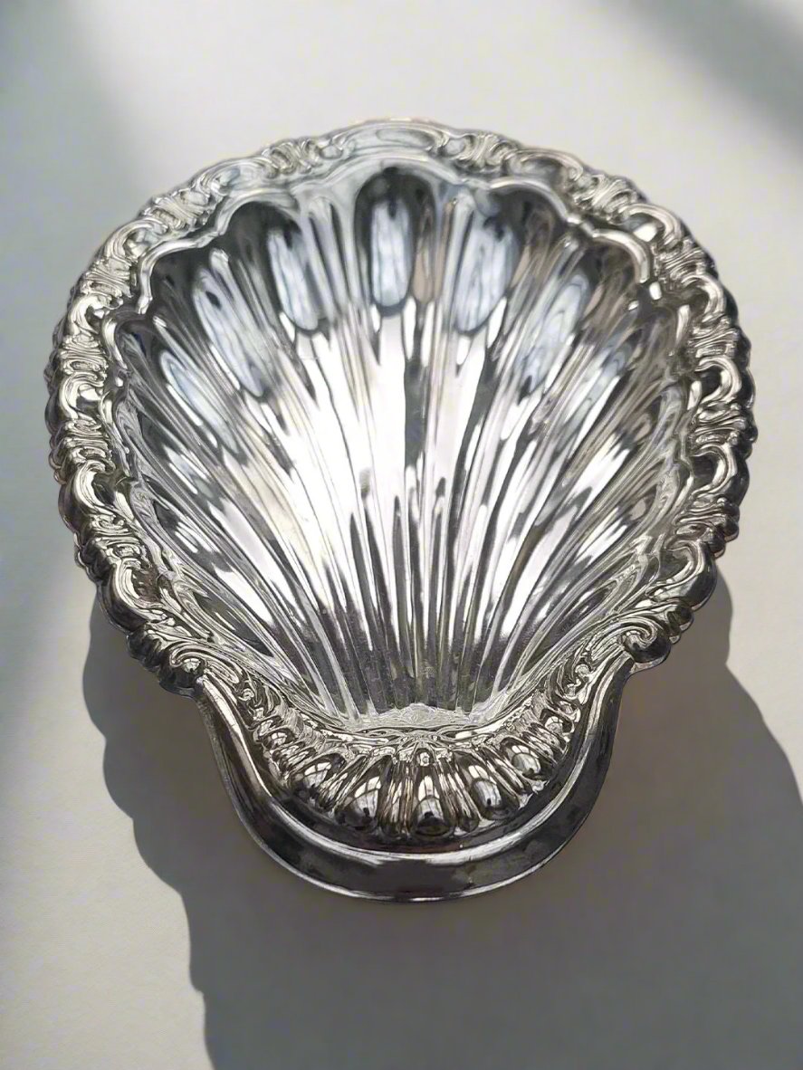 The Medium Shell Vide Poche by Les Objoies is a detailed, silver-plated bowl shaped like a scallop, resting on a smooth, light surface. Its intricate, embossed design features delicate details along the edges that reflect light to accentuate its polished finish, making it an exquisite vide poche.