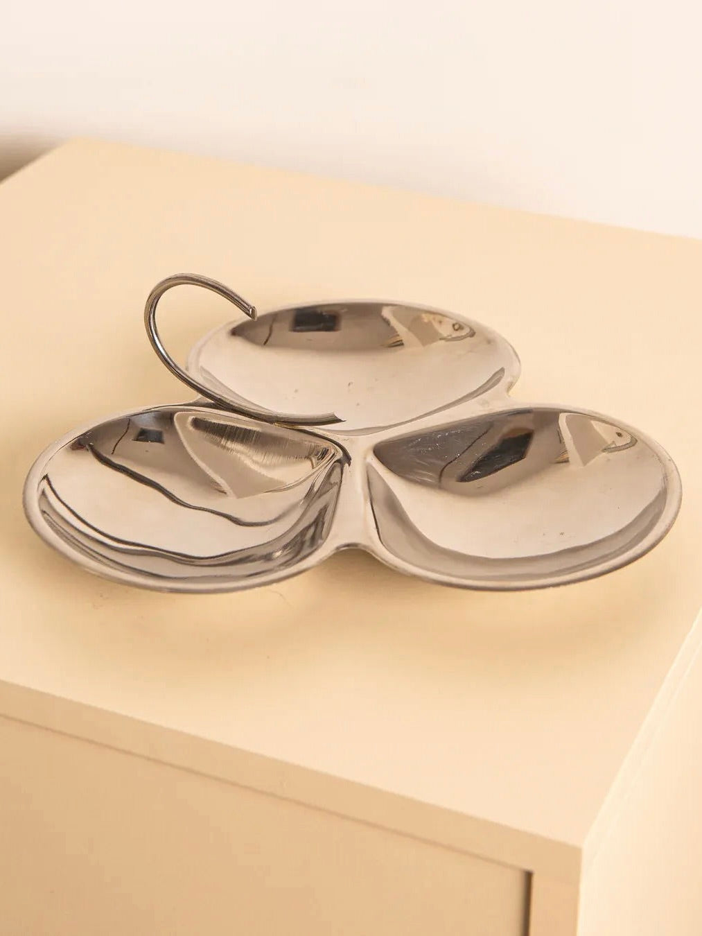 The "Cloverleaf" Shaped Vide-poches 80's by Treaptyque, a shiny, stainless steel serving tray with three sections and a handle, sits on a light wooden surface. Its reflective design captures ambient light, blending modern minimalism with an 80s vibe.