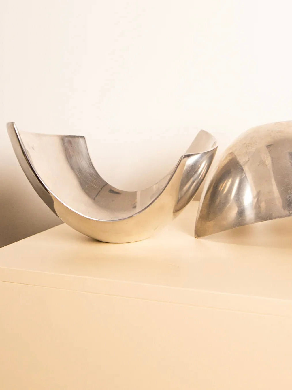 Treaptyque's Pair of "U"-shaped Fruit Baskets 80's are minimalist metal sculptures displayed on a light-colored surface against a plain wall. These retro-inspired pieces feature smooth, curved shapes crafted from silver-plated metal with a reflective finish.