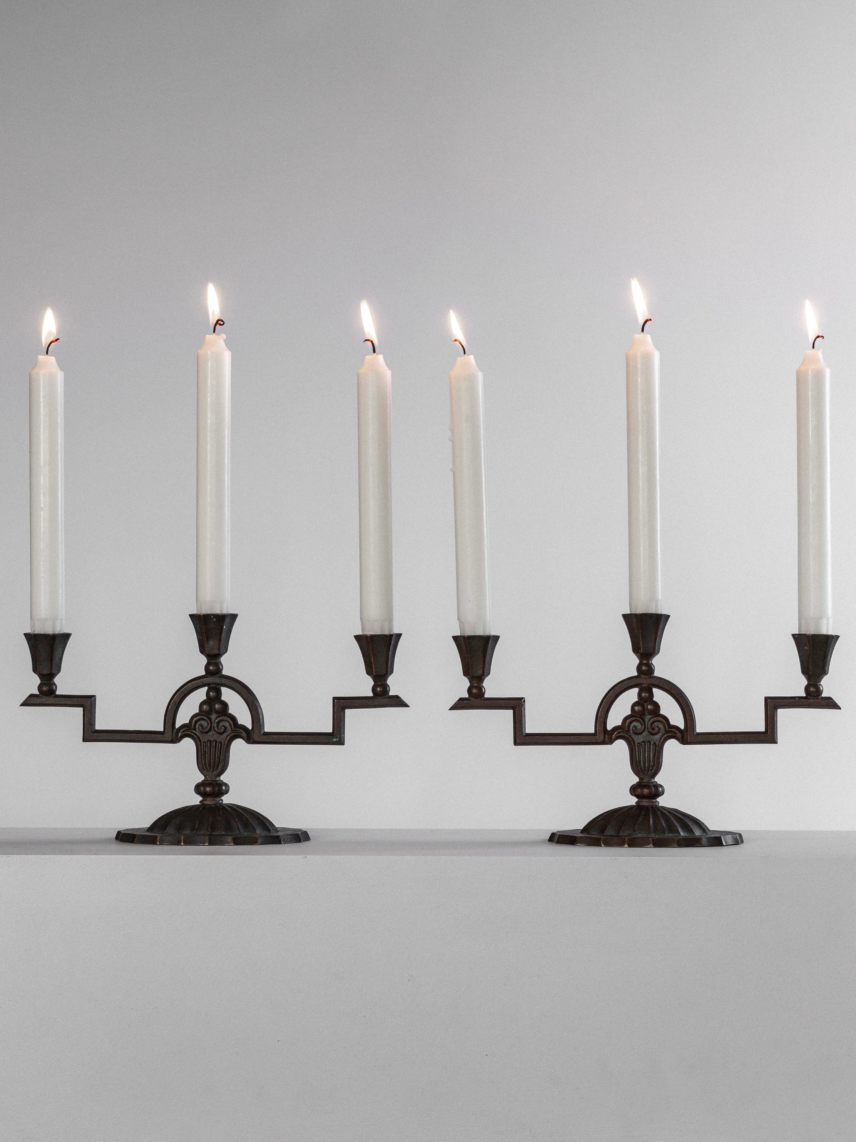 A pair of Art Deco Bronze Candelabras from the 1930s by Spigel, each holding three tall lit white candles. These elegant pieces boast an ornate design with curved arms and a decorative base, set against a plain white background. A true nod to Nordiska Juvelaktiebolaget craftsmanship.
