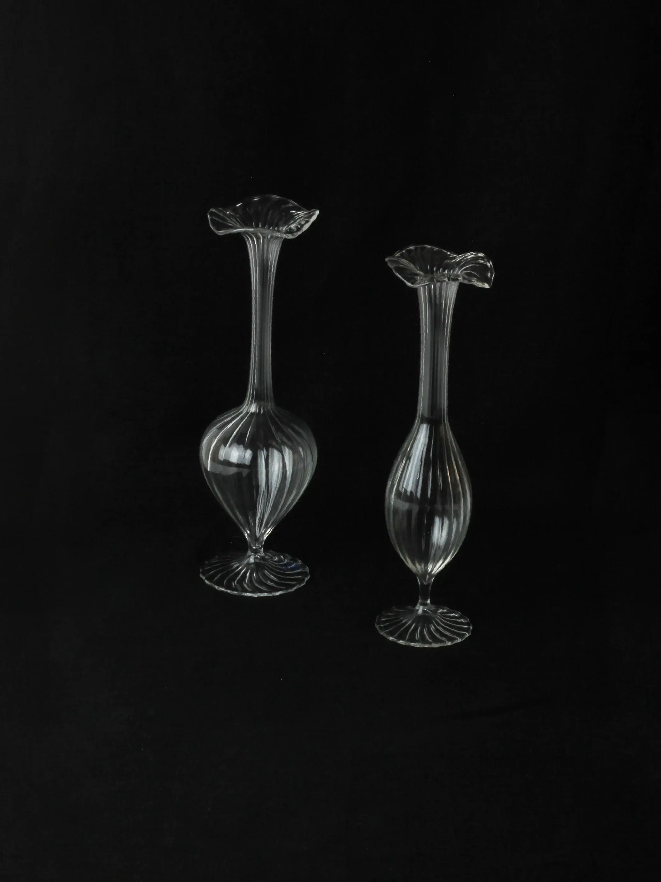 Two elegant, clear Murano candle holders from Boga Avante Shop, featuring elongated necks and flared rims, are showcased against a black background. The vintage blown glass design includes ribbed patterns and robust round bases, offering a classic, timeless style.