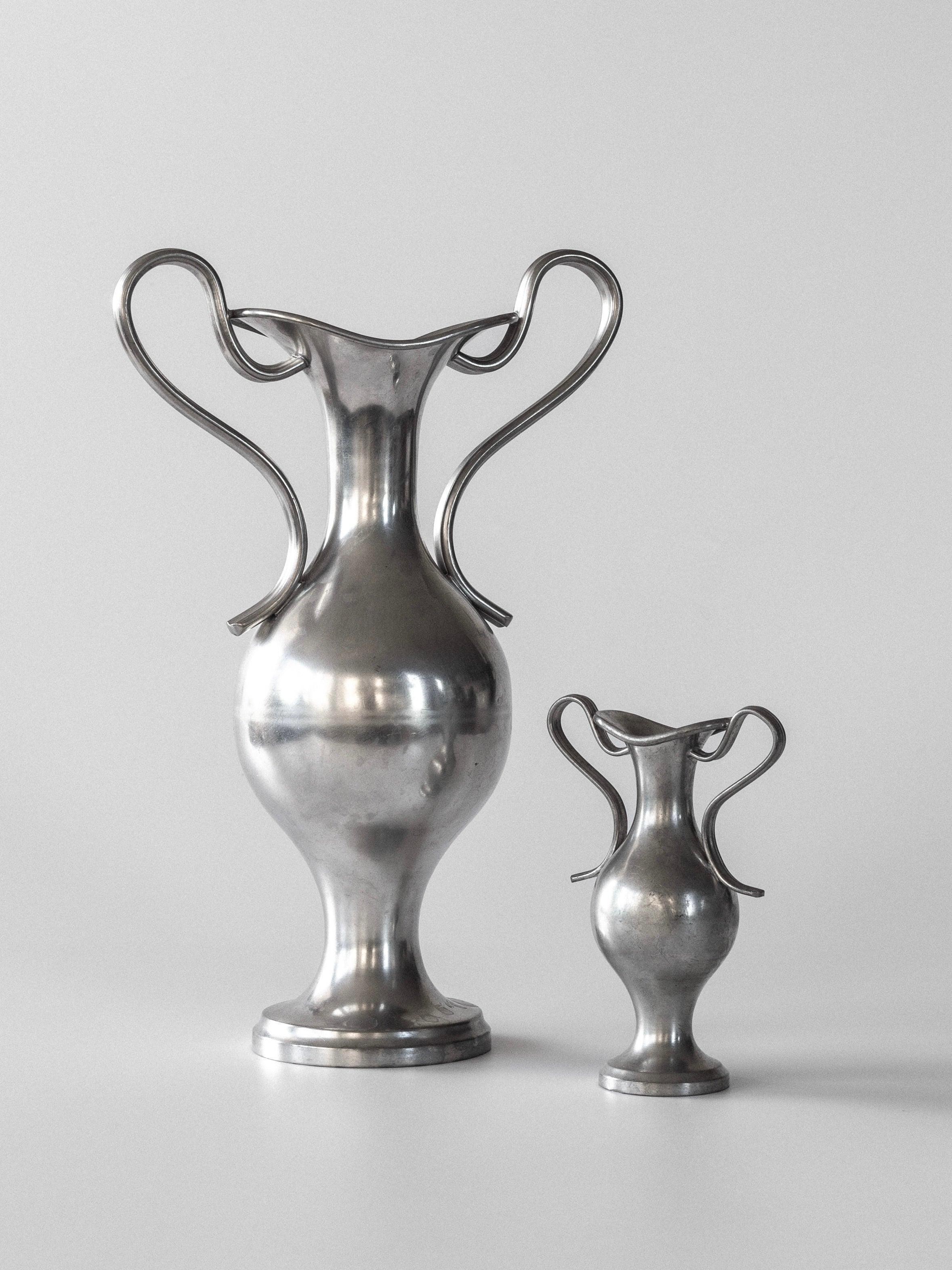 Displayed against a plain backdrop, the Pair of Neoclassical Metal Vases by Spigel from 1953, Sweden, features ornate handles. The taller vase is on the left and the shorter on the right, both showcasing smooth, reflective surfaces and elegant curvy designs.