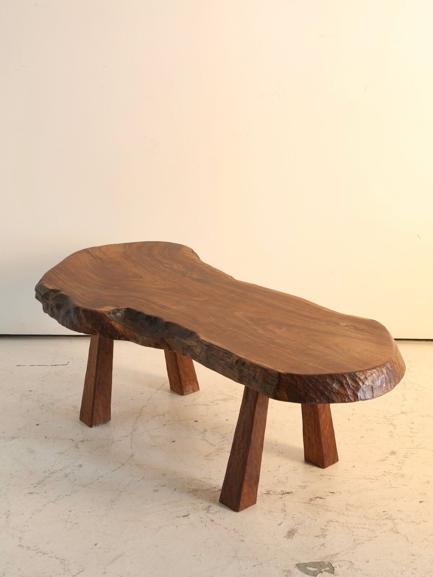 The 'French Folk' Coffee Table by Ancien et Jolie boasts a 1970s-inspired rustic waney edge, with three angled legs and a polished surface. Its adze carved details stand out against a plain, light-colored wall, evoking the natural charm of vintage France.