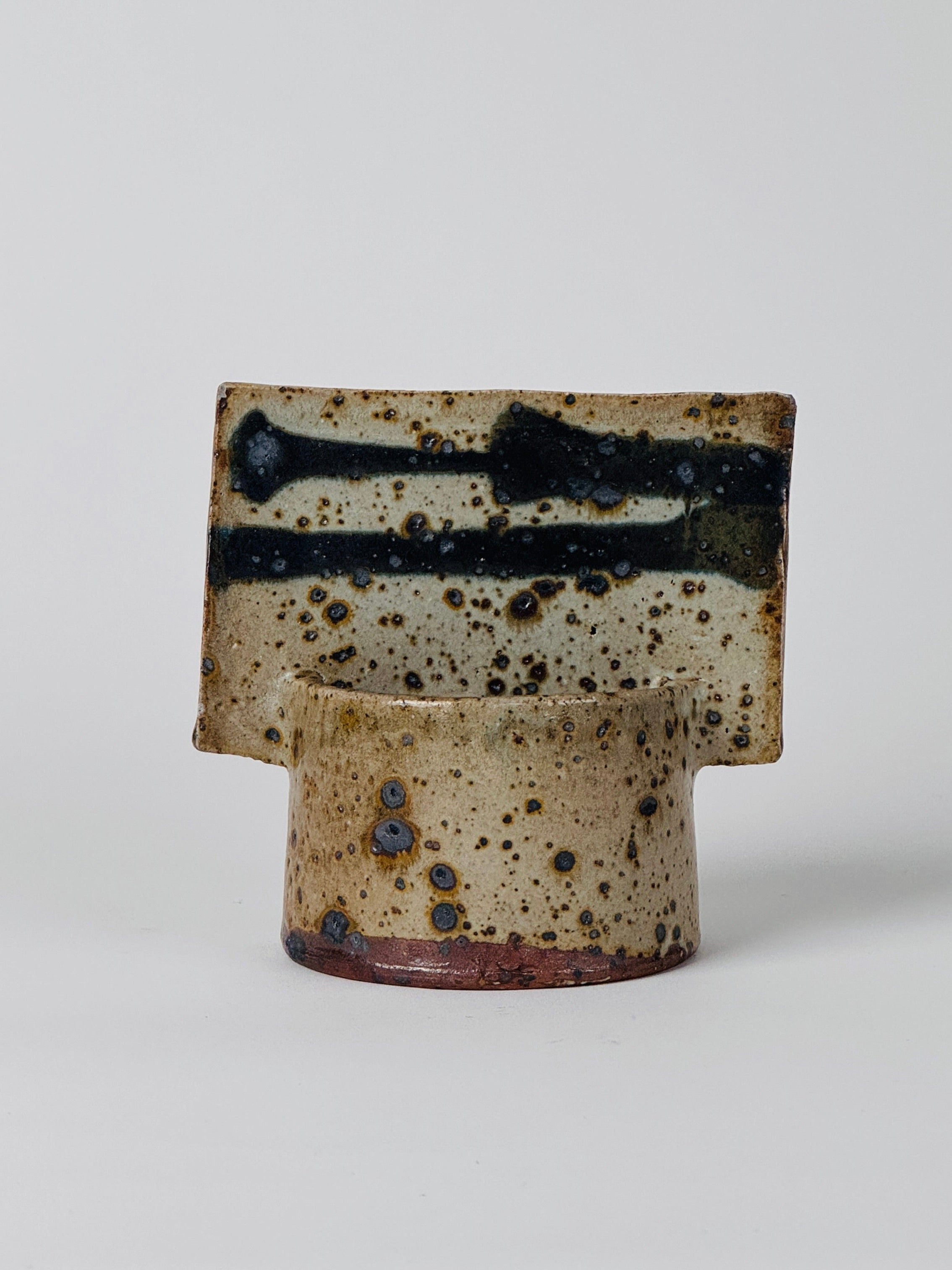 The Geometric Vase by Gustave Tiffoche from Figuresdesign features a cylindrical ceramic base paired with a distinctive, flat, rectangular top that evokes the essence of pyrite stoneware. Its speckled brown and beige surface with dark blue streaks and spots imparts a rustic, artisanal appearance. The vase stands against a plain white background.