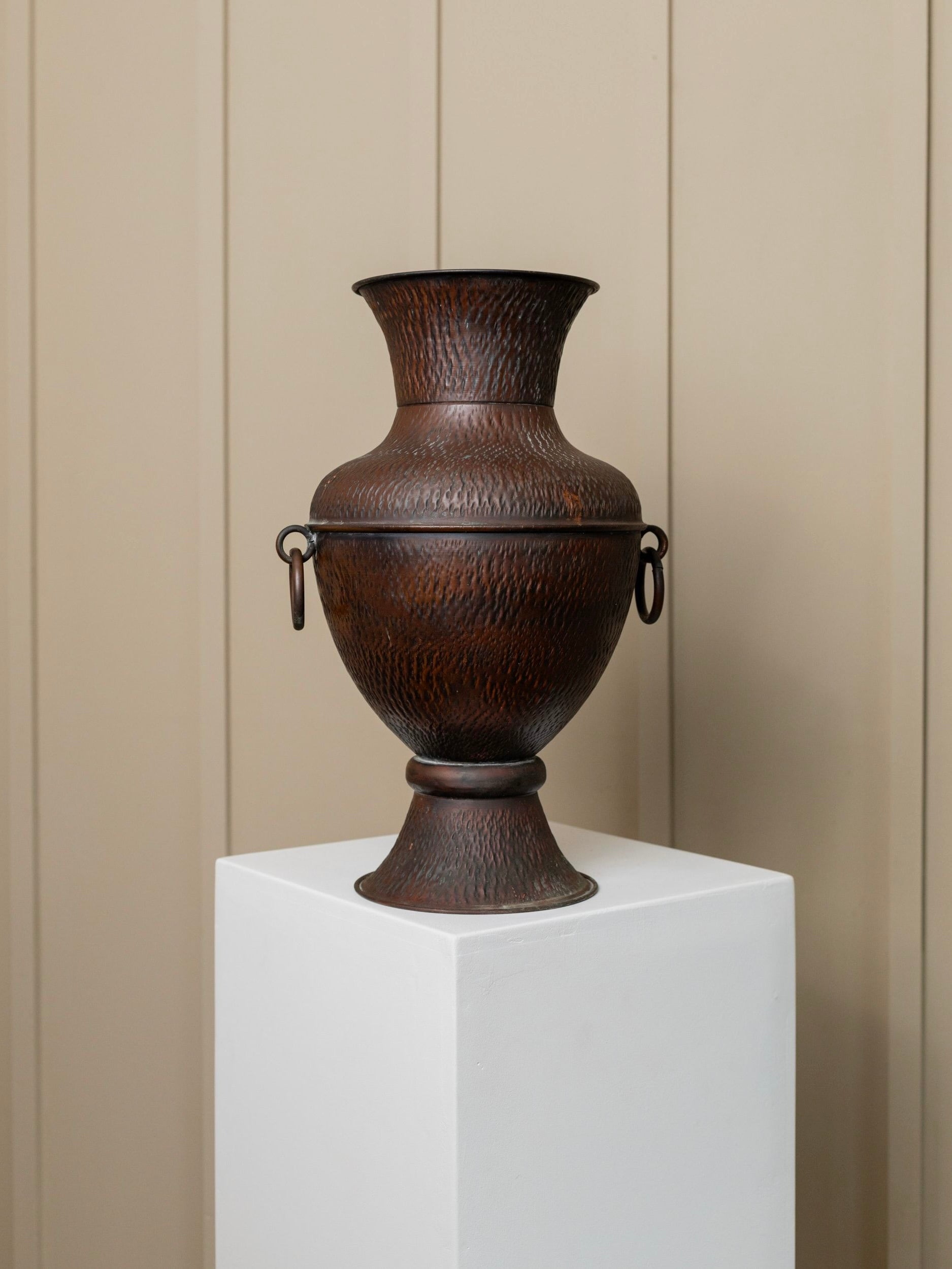 Display: A Vintage Copper Jug from Bottega Jacobs, made in Italy in the 1960s, featuring a textured surface and a wide flared opening. Its intricate patterns and dark bronze color enhance its elegant presence as it rests on a white pedestal against a light beige, vertically paneled backdrop. This decorative piece is adorned with two small handles on either side, adding to its charm.