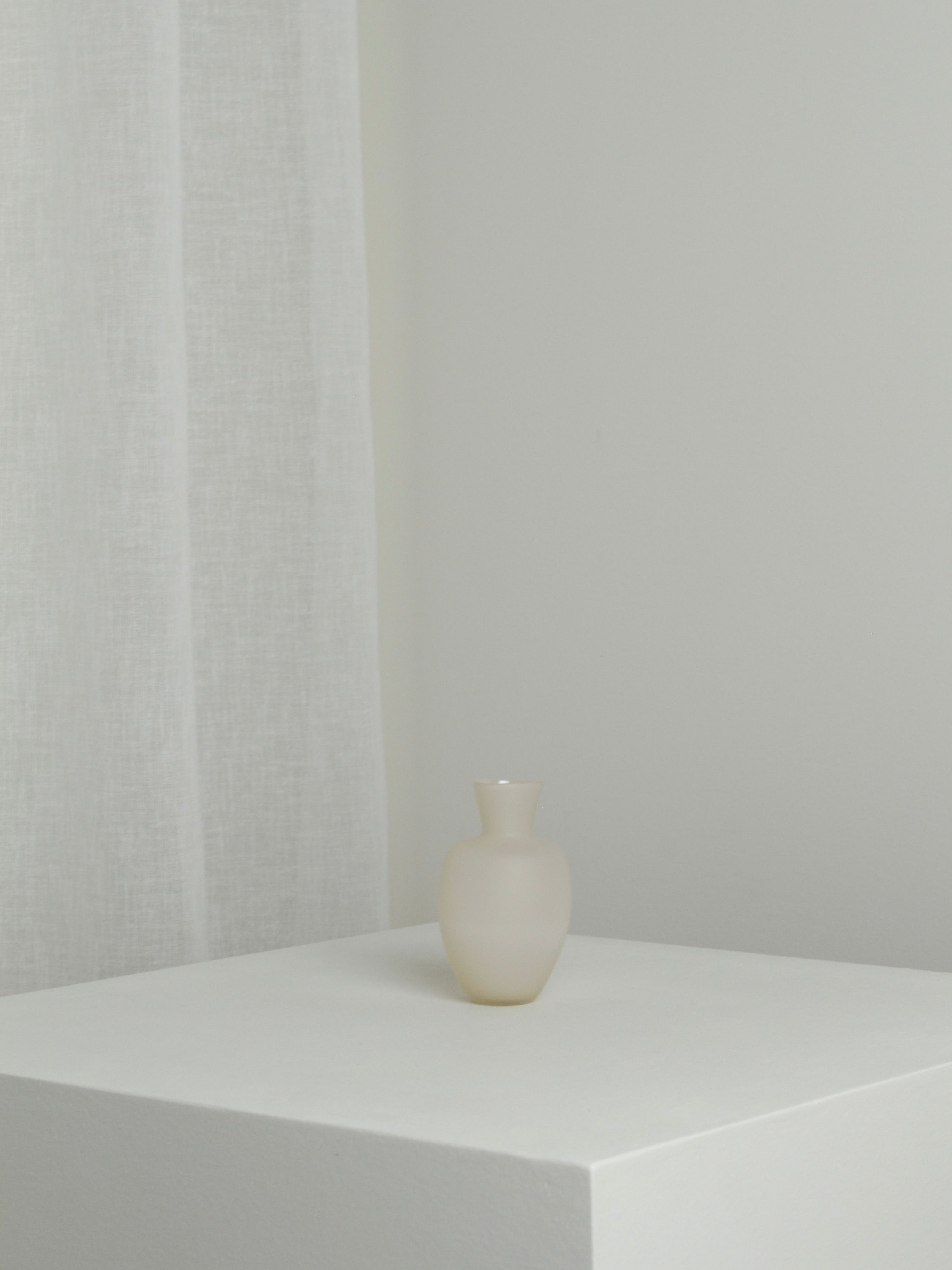 A small, mouth-blown vase from Verre d'Onge's "Small Vase in Moon 2" collection is elegantly placed on a white pedestal against a light gray background. A sheer white curtain is partially visible in the left corner of the image. The minimalist and serene scene is bathed in soft moonlight.