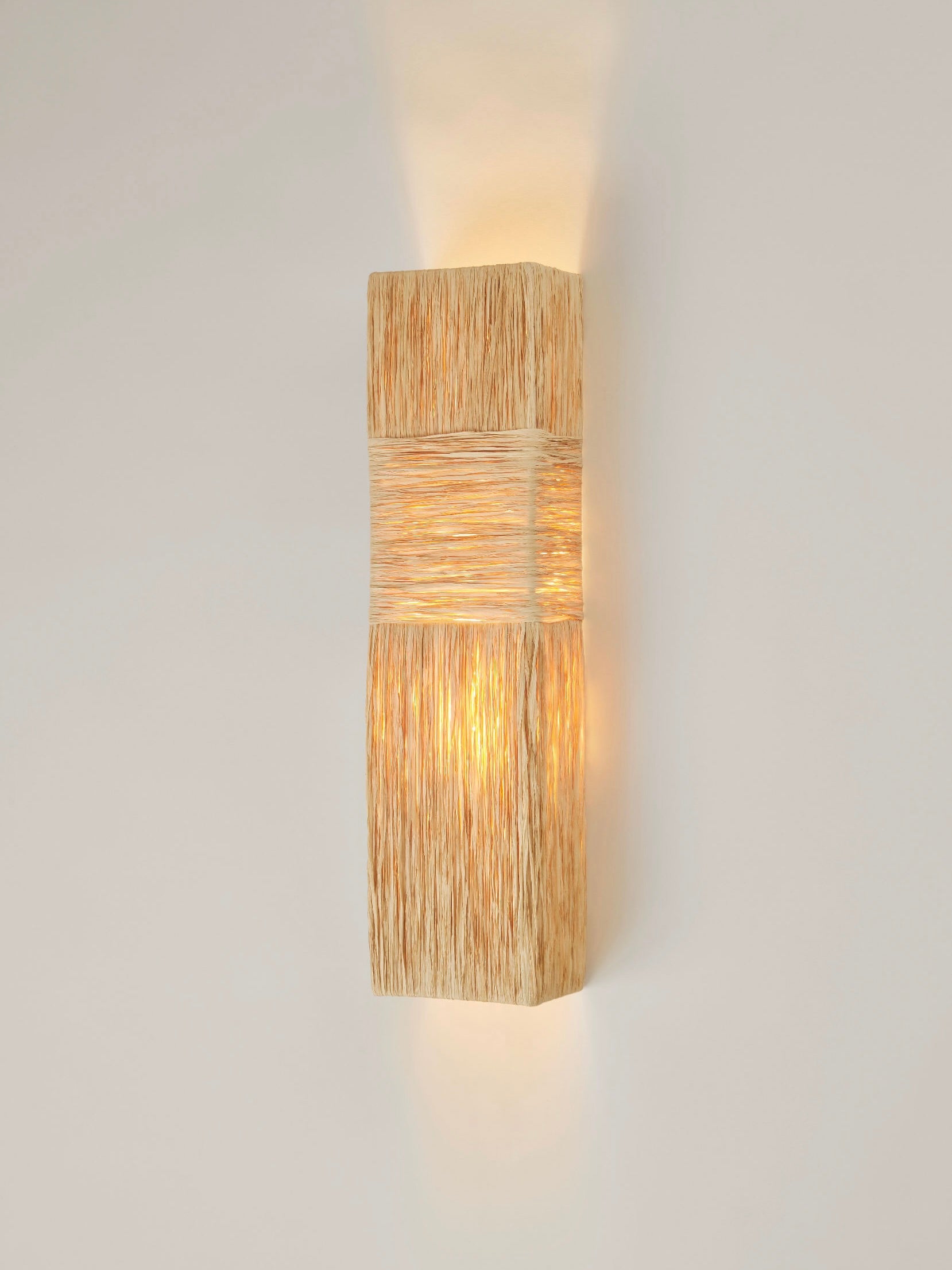 The "Eliane" Wall Sconce by HAUVETTE & MADANI features a woven rectangular design that emits a warm, soft light. Its natural earthy tones evoke 1930s dinners and create a cozy ambiance against white walls, adding elegance reminiscent of Hauvette Madani furniture.