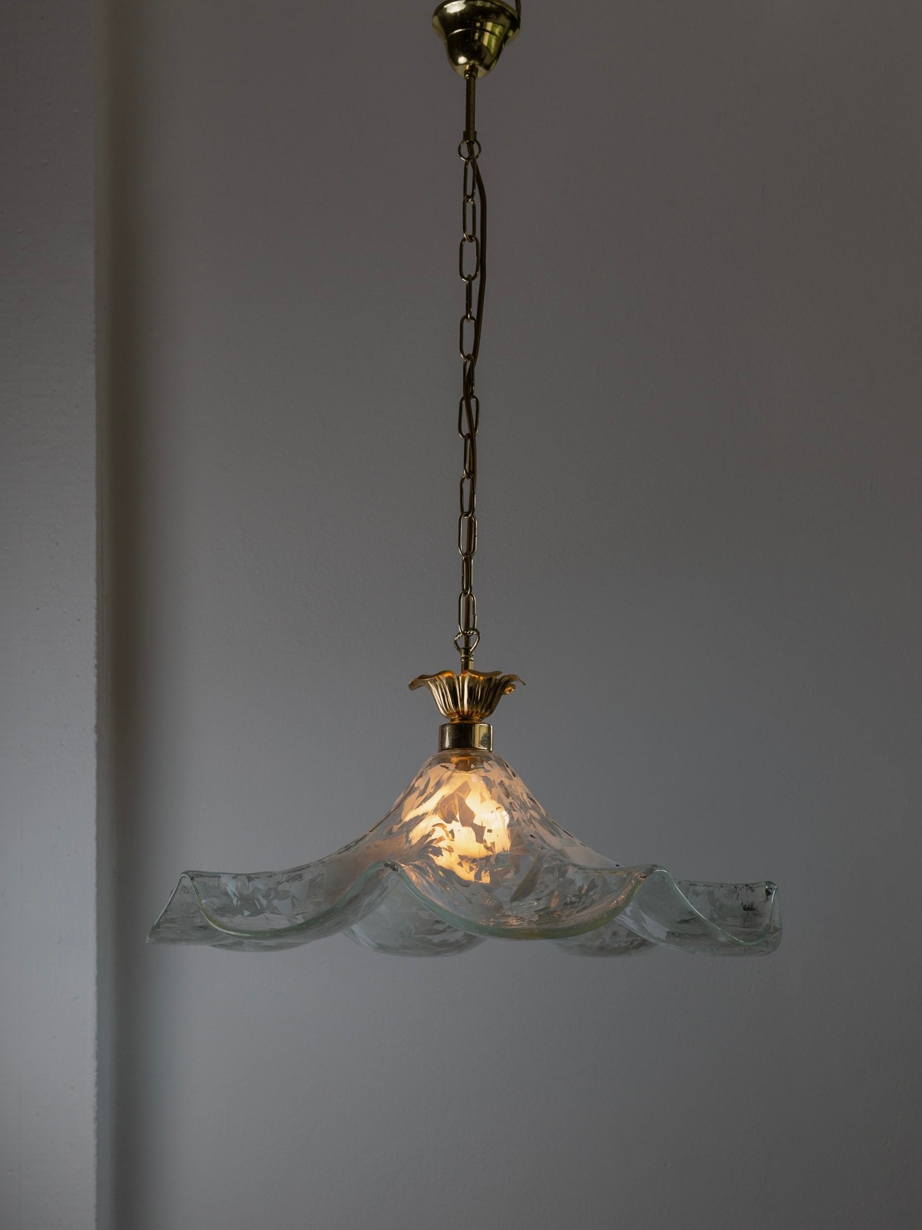 A vintage ceiling lamp crafted by Bottega Jacobs, featuring a gold chain and an ornate flower-shaped Murano glass shade from the 70s in Italy. The wavy and translucent decorative glass emits a warm glow against a plain background.