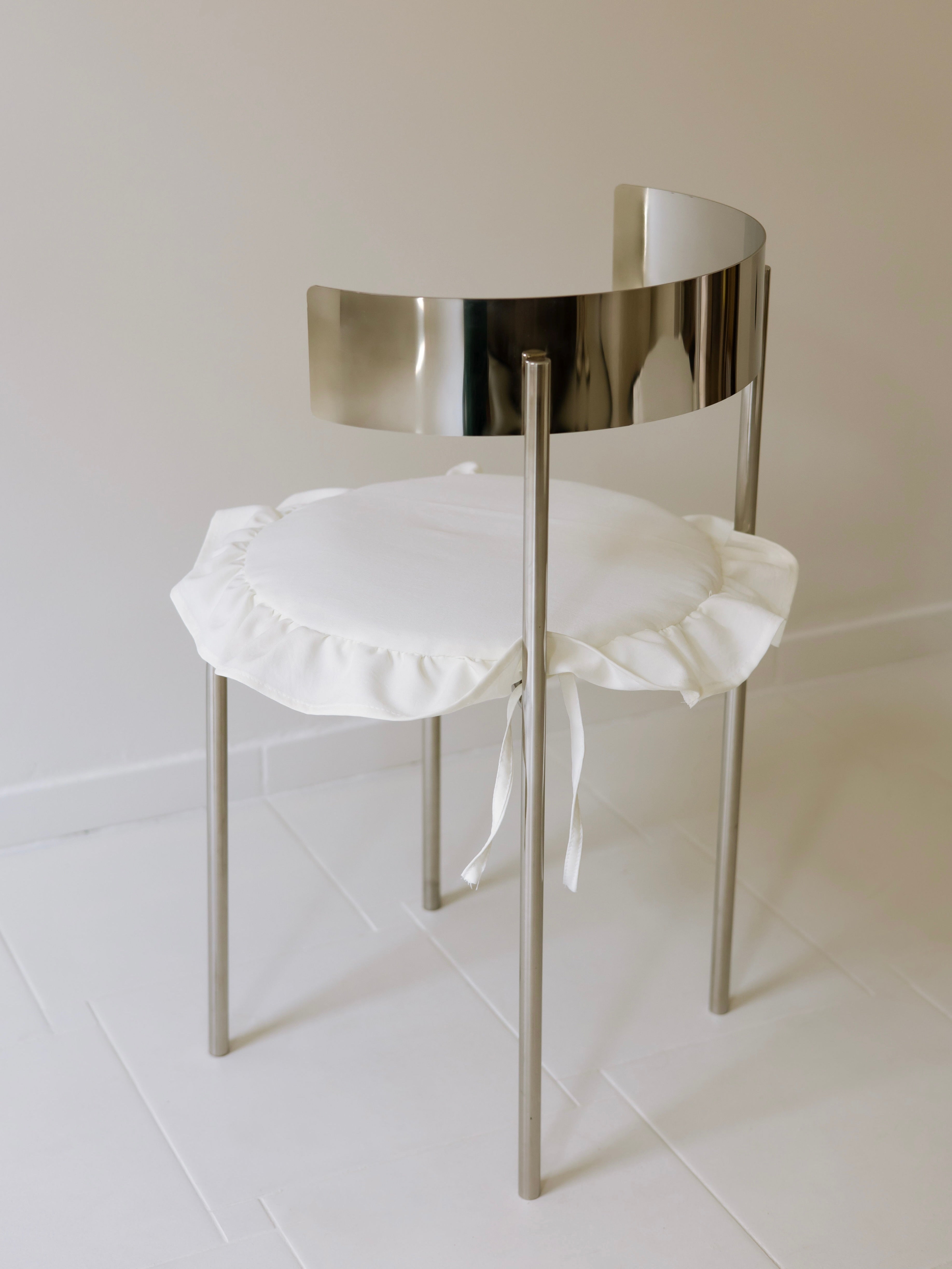 The Good Living&Co. "Meadow" Chair - Skirted features a stainless steel frame with a reflective backrest and a round cotton-cushioned seat cover tied with ribbons. Set on light tiles and against a neutral wall, it's stylish and sophisticated.