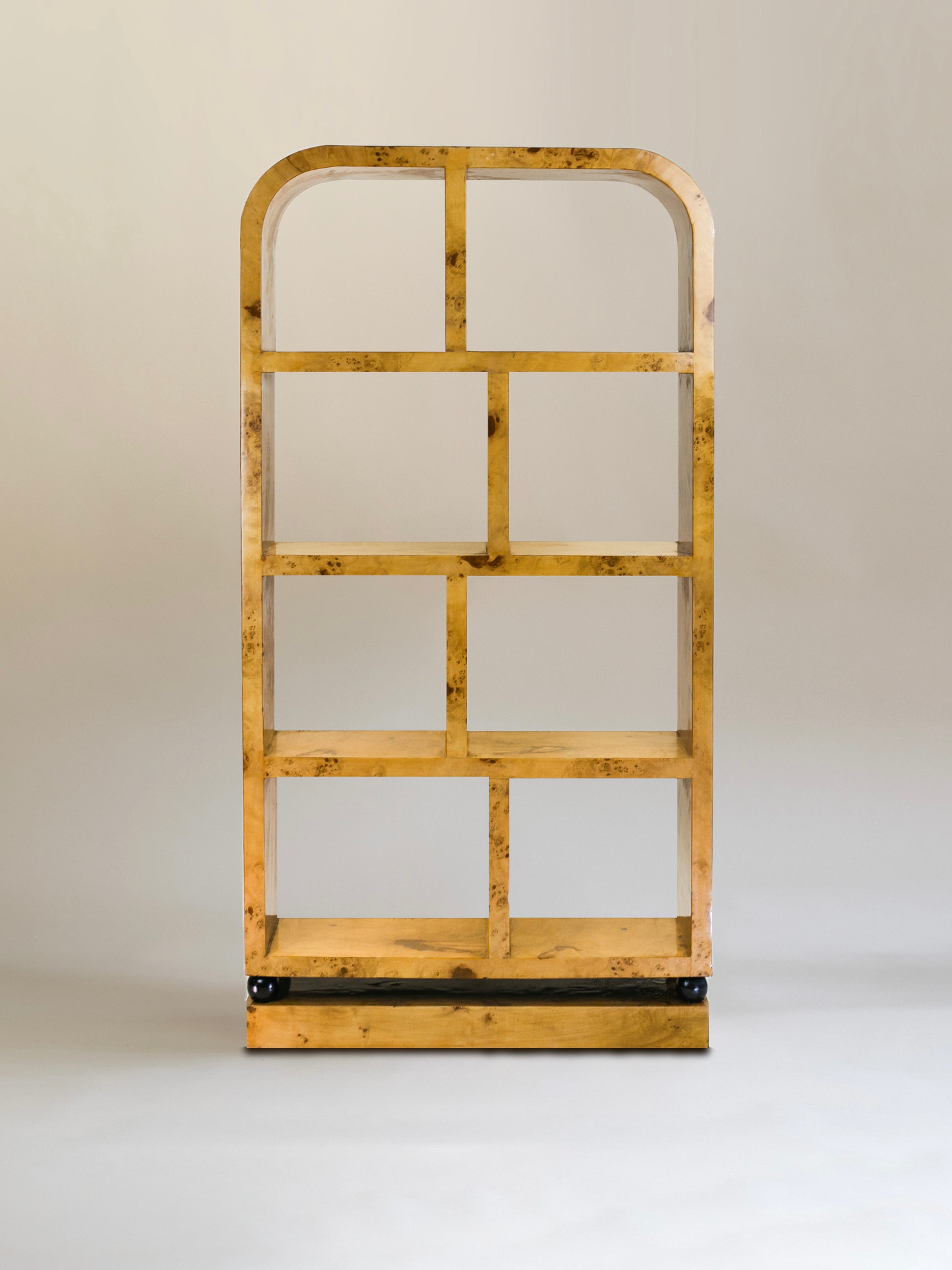The Calisto Double Bookcase by Meola Interiors features a minimalist design with a wooden, open shelf unit. Its rectangular frame and arched top form six evenly spaced compartments on a light gray backdrop, while the natural, warm finish adds mid-century elegance.