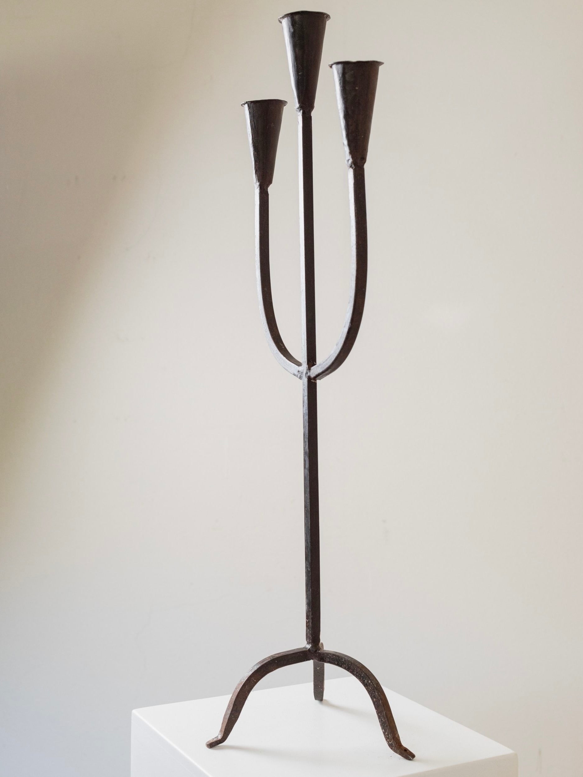 An XXL Brutalist metal Candle Holder from the 1960s by Bottega Jacobs stands on a white pedestal against a plain beige background. Its minimalist black iron design with slender arms curving upwards evokes the timeless beauty of an Italian candle holder.