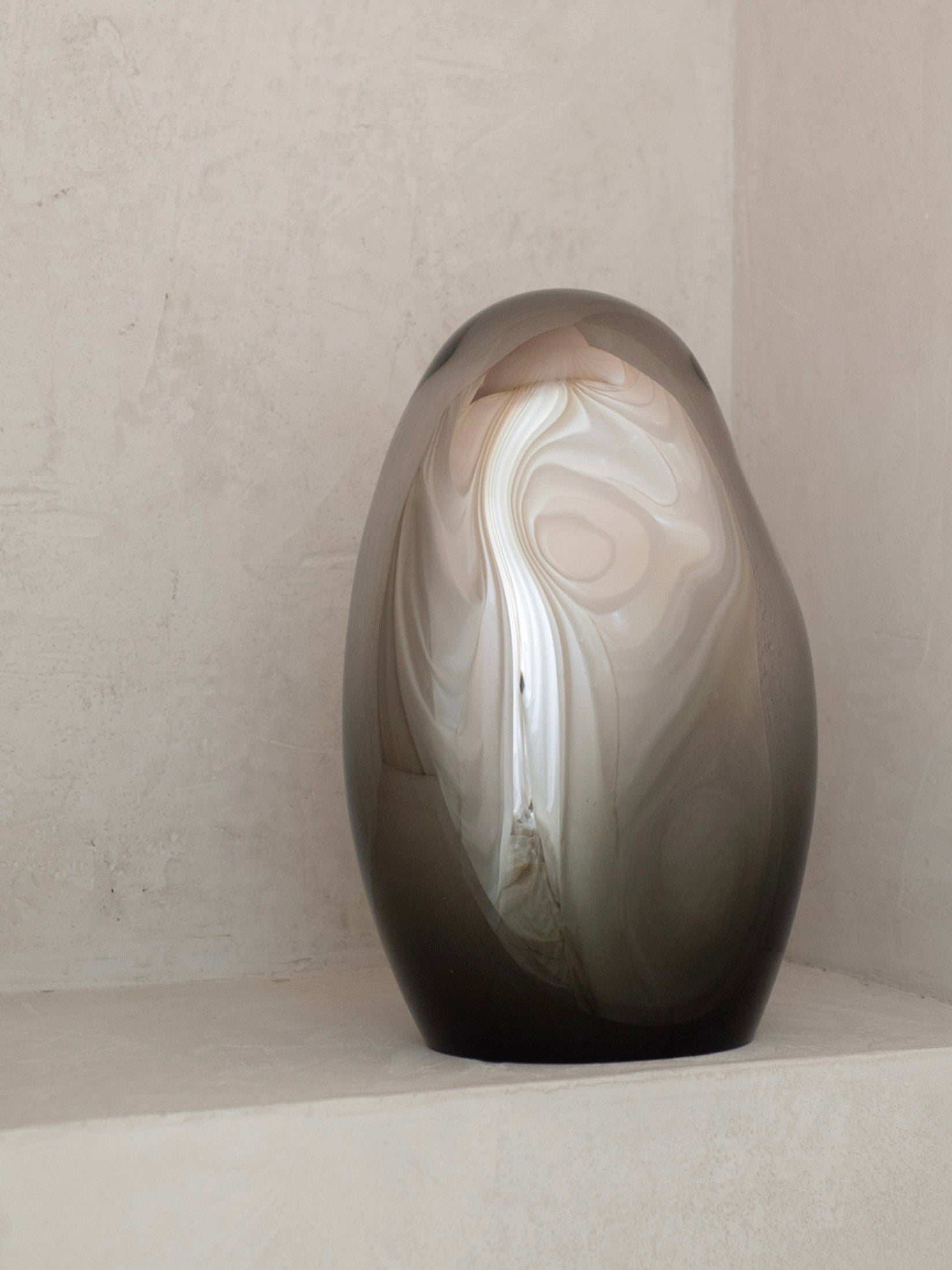 A No. 06 Standing Mirror Sculpture - Smoke by STUDIO MILENA KLING, crafted from mouth-blown glass with a swirled pattern of whites, browns, and grays, is showcased on a light-textured surface against a neutral wall.