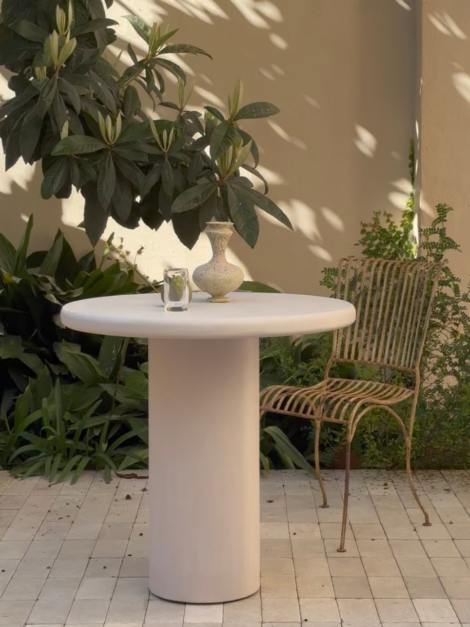 A handmade Yay Dining Table by MarlotBaus, featuring a minimalist design, sits on a greenery-filled patio. On the table are a ceramic vase and glass. A metal chair with a woven pattern is positioned beside it, showcasing craftsmanship as sunlight casts shadows on the tiles.