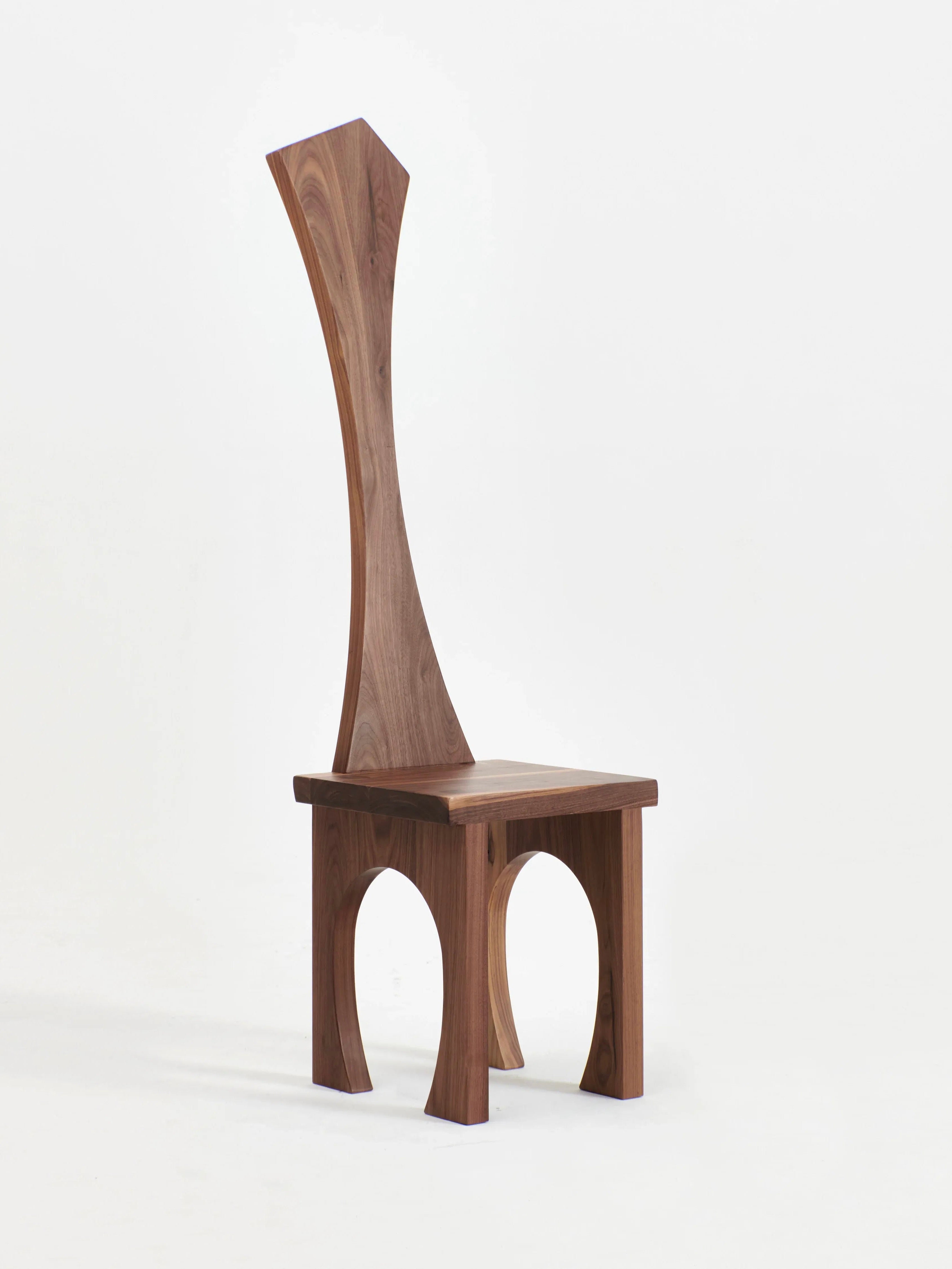 A modern wooden chair with an artistic design, the Portugal Chair No 8 by Project 213A boasts a high backrest that curves inward. Its seat is supported by four legs, each featuring arched cutouts. Hand carved from solid walnut, the sleek natural wood finish enhances its contemporary and unique appearance.