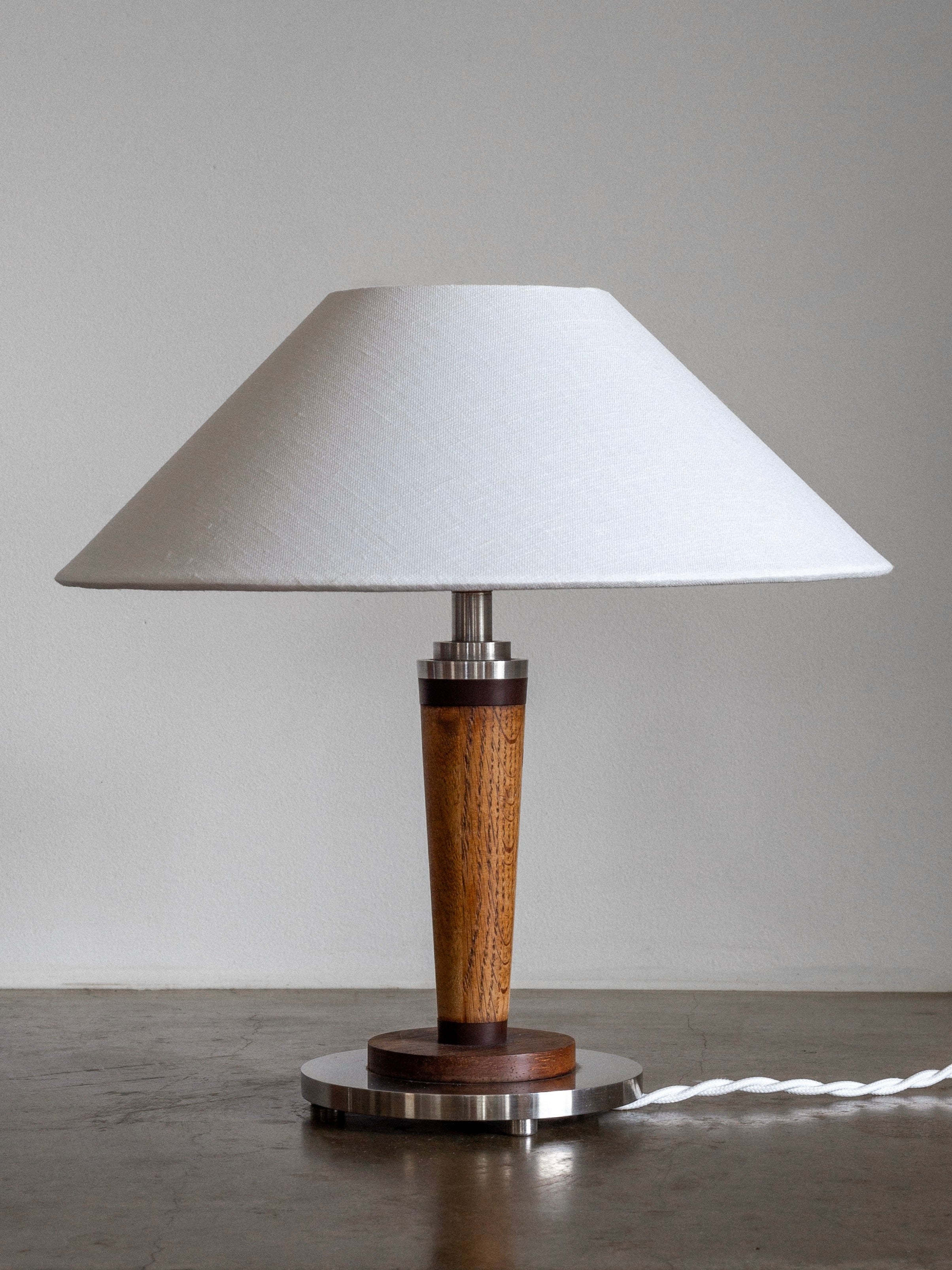 The Spigel Mid-Century Metal and Wood Lamp, featuring a conical white lampshade and a teak wood and metal base with a twisted white cord, is placed on a smooth, gray surface against a plain gray wall.