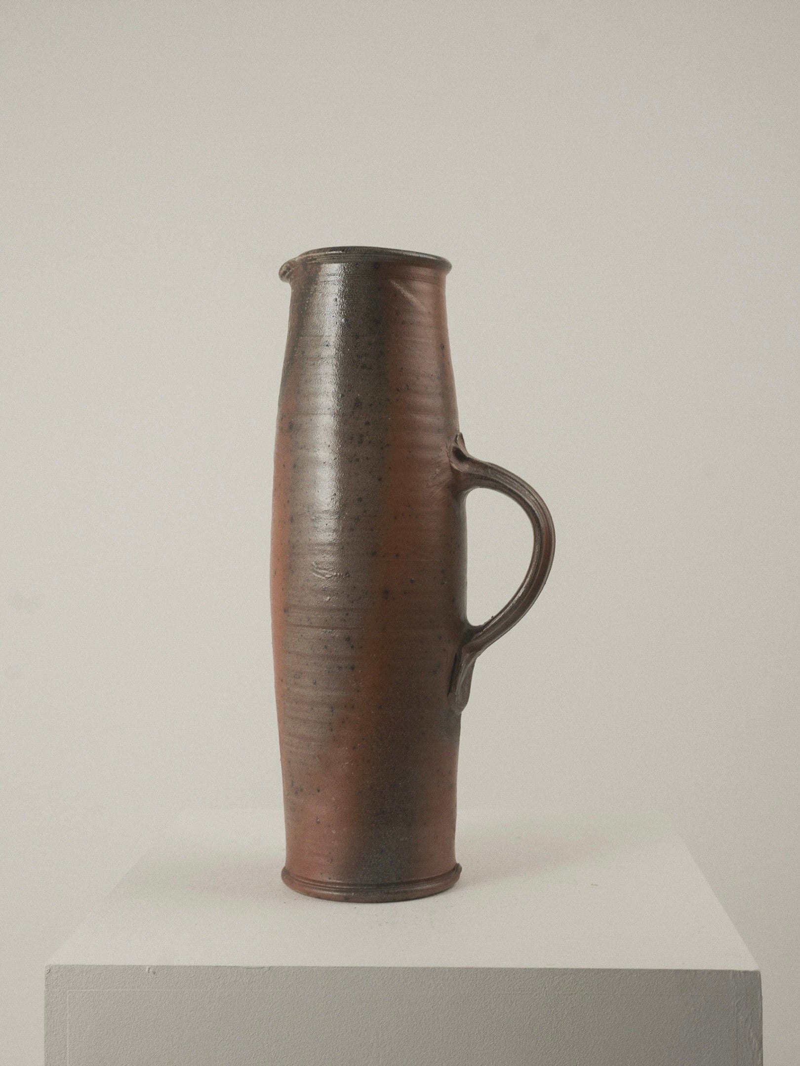 The Large Pitcher by Gustave Tiffoche from Figuresdesign, featuring a tall, slender stoneware form with a warm brown finish and subtle texture, sits on a white pedestal. Its single curved handle and small spout offer minimalist elegance reminiscent of Tiffoche against a plain background.