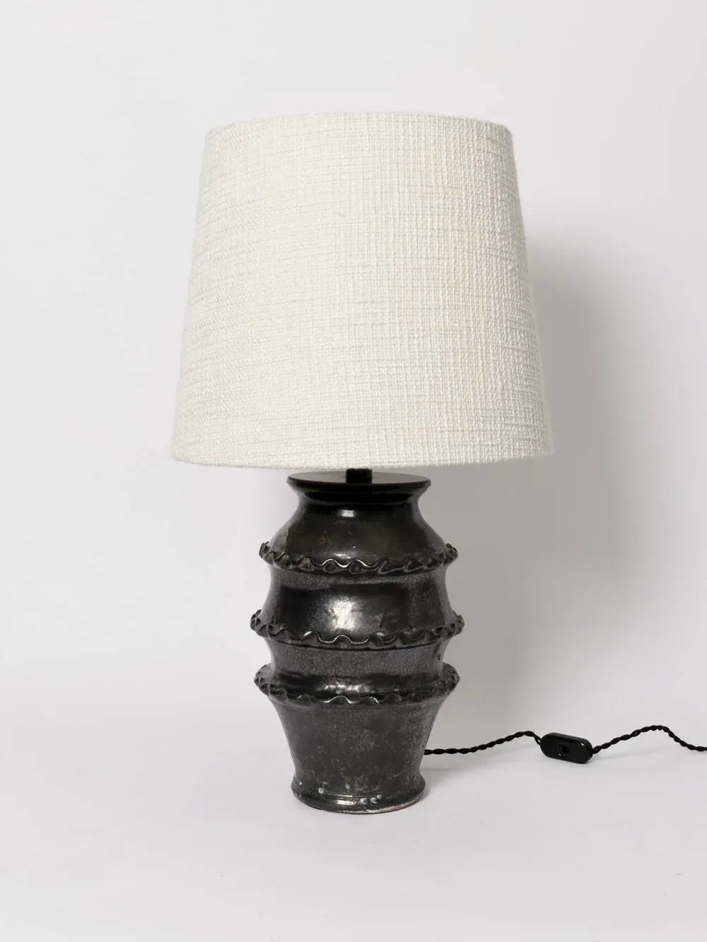 The Black Glazed Ceramic Table Lamp from Barracuda Interiors features a textured white shade atop an ornate dark metal base accented with ribbed and wavy patterns. The design is elegantly completed with a black glazed finish and includes a discreet black power cord for seamless integration.