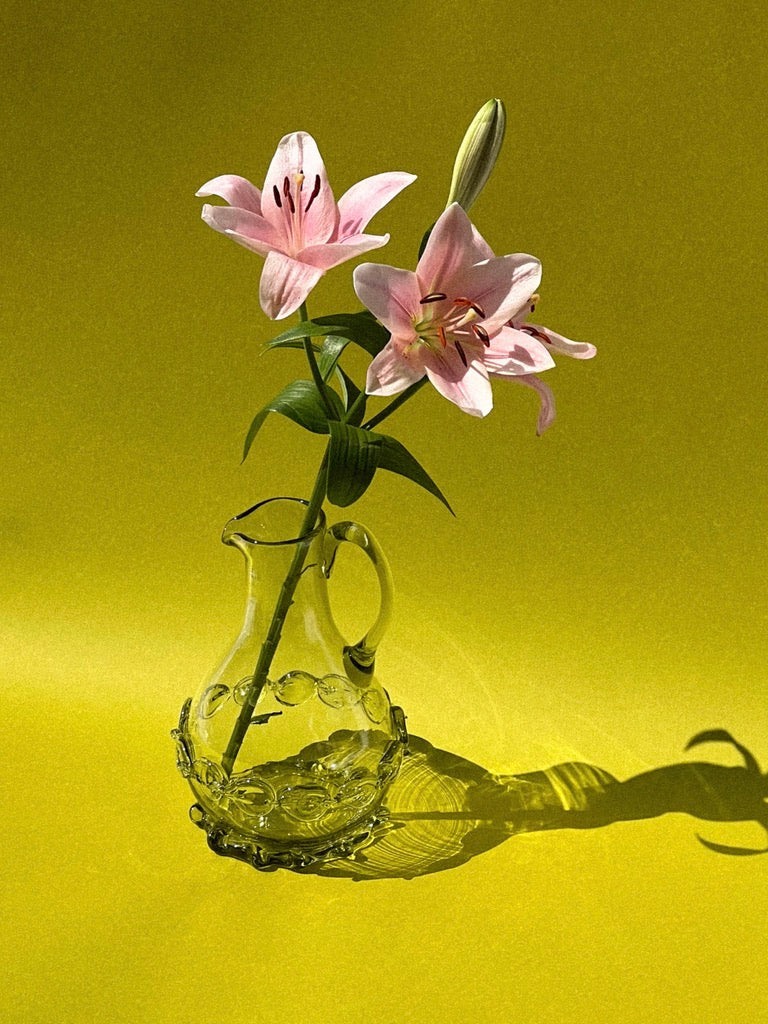 A clear, mouthblown carafe from Lucie Claudia Podrabska holds two blooming pink lilies against a solid yellow background. The flowers cast distinct shadows, creating a vibrant and striking contrast with the backdrop. The composition highlights the delicate details of the lilies and the artisanal craftsmanship of the carafe.