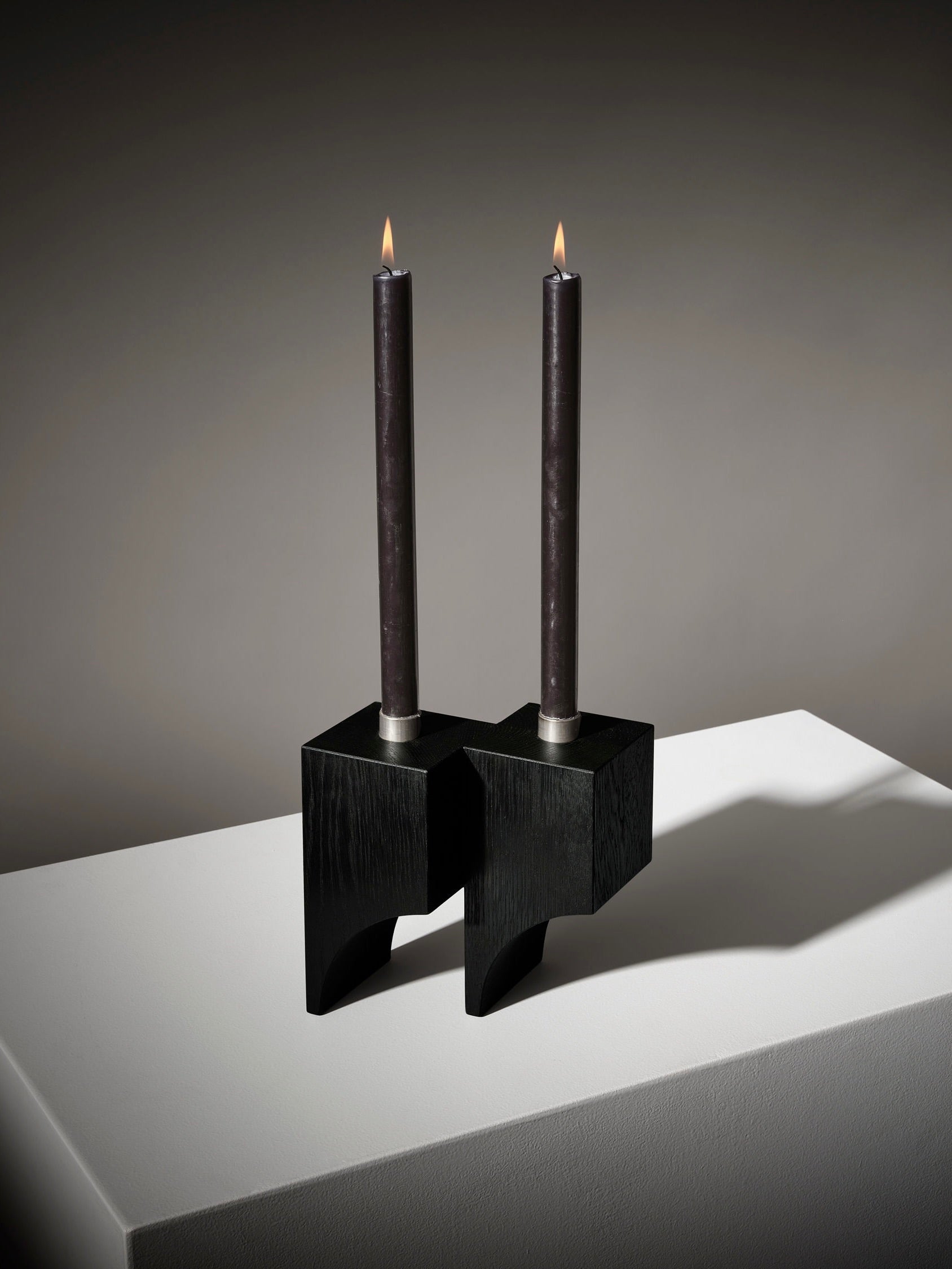 Two lit black candles in a sleek Acer Candle Holder R:2 by Mokko rest on a white surface, with a gradient backdrop from light to dark gray, emphasizing its minimalist design.