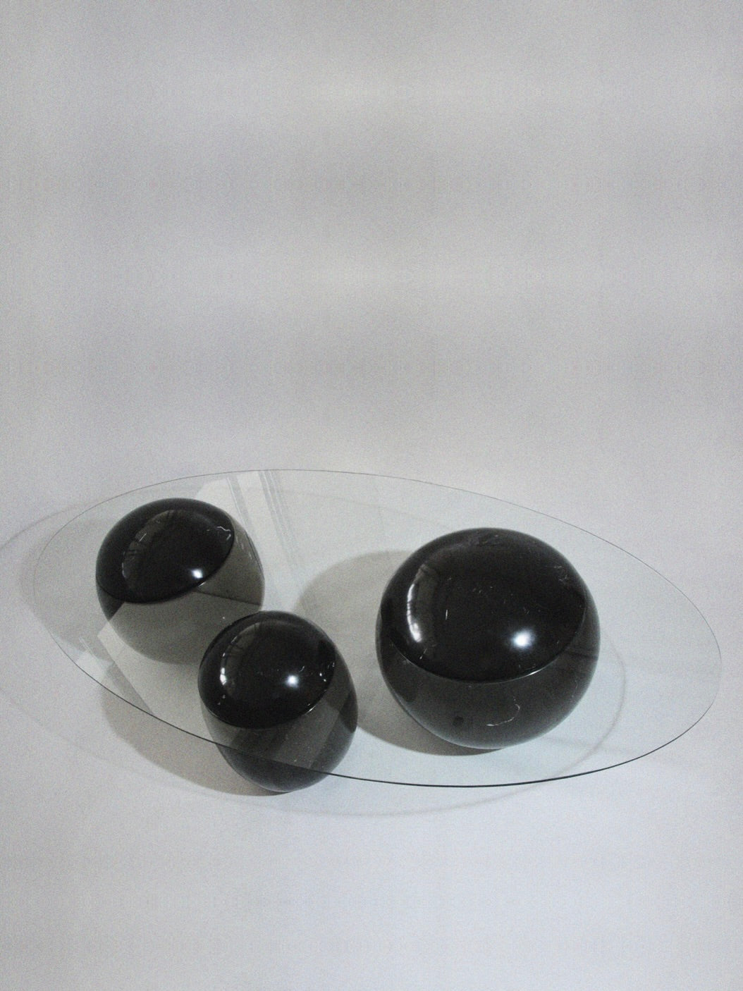 Presenting Panorammma's 'Ovni Uovo' Coffee Table: a minimalist design masterpiece with a clear oval glass top gracefully perched on retro-futurist black polished stone spheres of various sizes, all set against a light gray background.