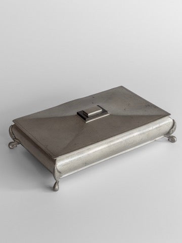 A rectangular metal box with a hinged lid and a small handle rests on four curled legs, known as the Art Deco Metal Box by Spigel. This box features a polished pewter finish and minimalist design that captures the elegance of Scandinavian metalwork against a plain backdrop.