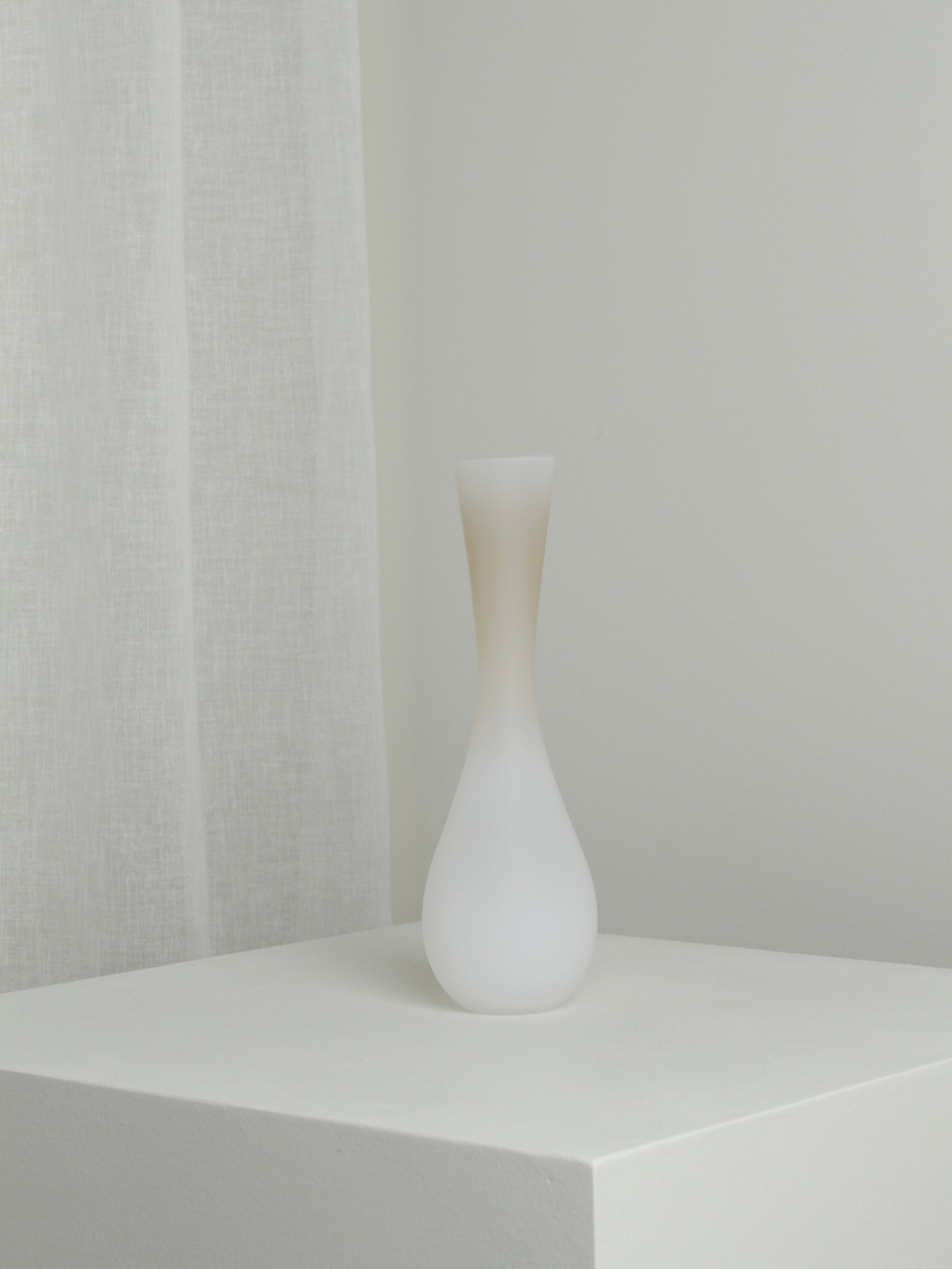 A minimalist scene featuring the Medium Vase in Mushroom from Verre d'Onge, a mouth-blown frosted glass piece with a narrow neck and wide base, placed on a white pedestal against a light background. A sheer white curtain is partially visible on the left side. The environment is neutral and calming.