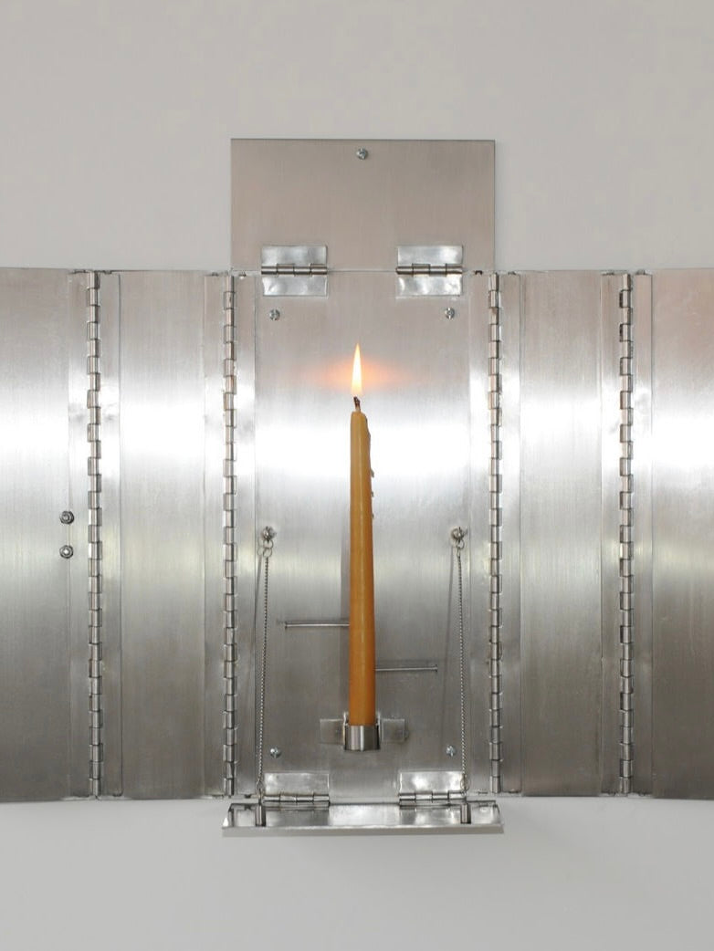 The 'Wax Clock' Wall Candleholder by Panorammma features a yellow candle in an exquisite holder on a silver, folding metal panel. Symmetrical hinges and a minimalist industrial design blend timeless charm with contemporary elegance as the flame softly illuminates the surface.
