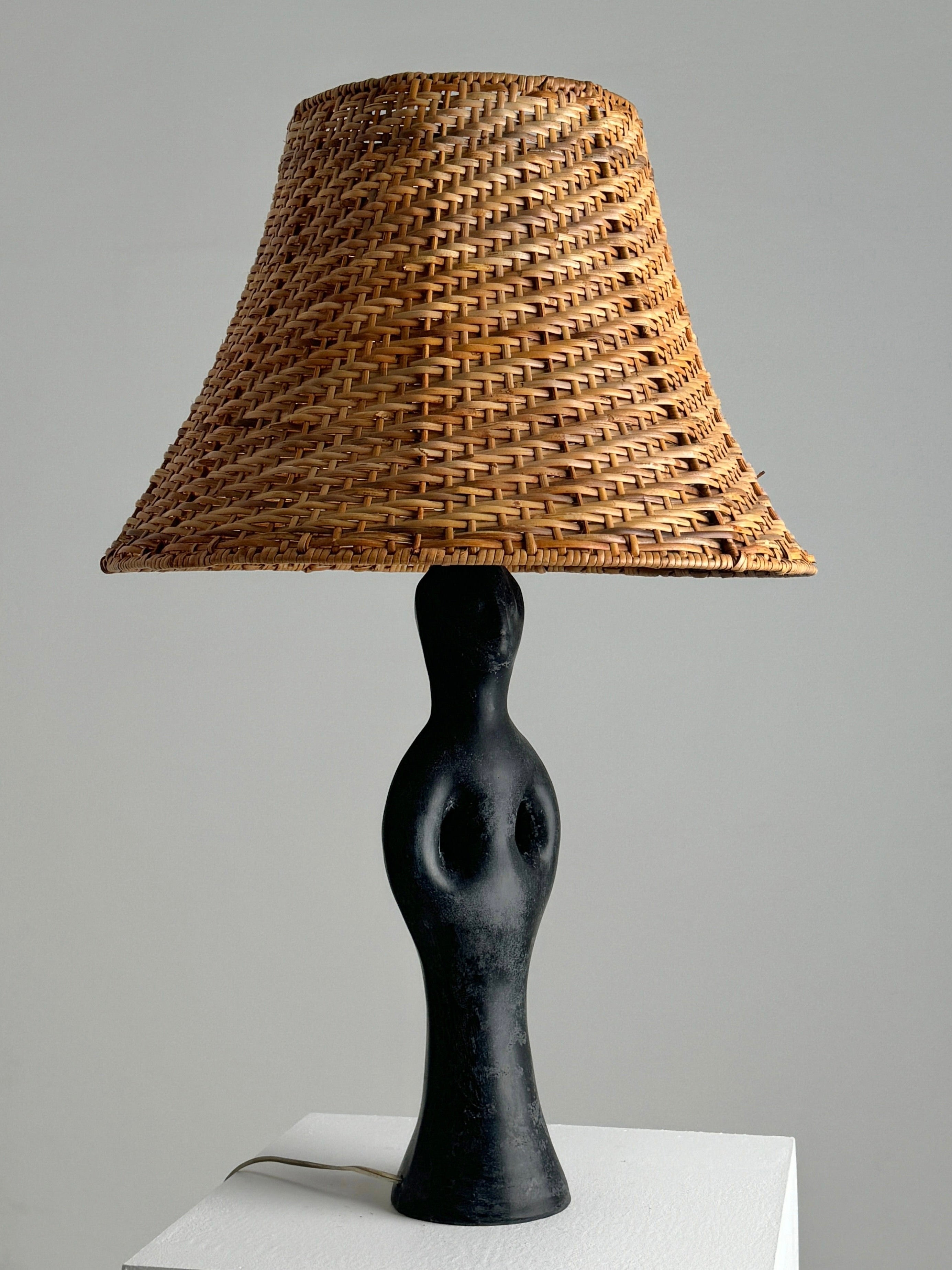 Figuresdesign's Black Marble Anthropomorphic Table Lamp features a sculpted black base resembling a human figure on a white surface. A naturally woven lampshade adds charm with its basket-like texture, evoking 1950s elegance and contrasting beautifully against the light gray wall behind it.