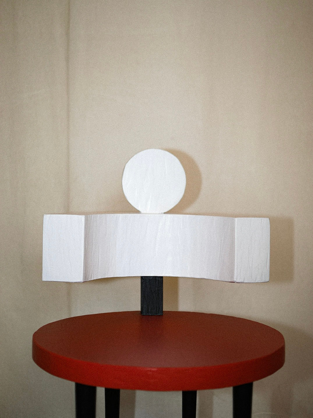 A modern textured white sculpture resembling a head and torso sits on Rooms Studio's Sculptural Chair II in Primary Colors, while a red circular table with black legs complements the scene against neutral beige curtains.