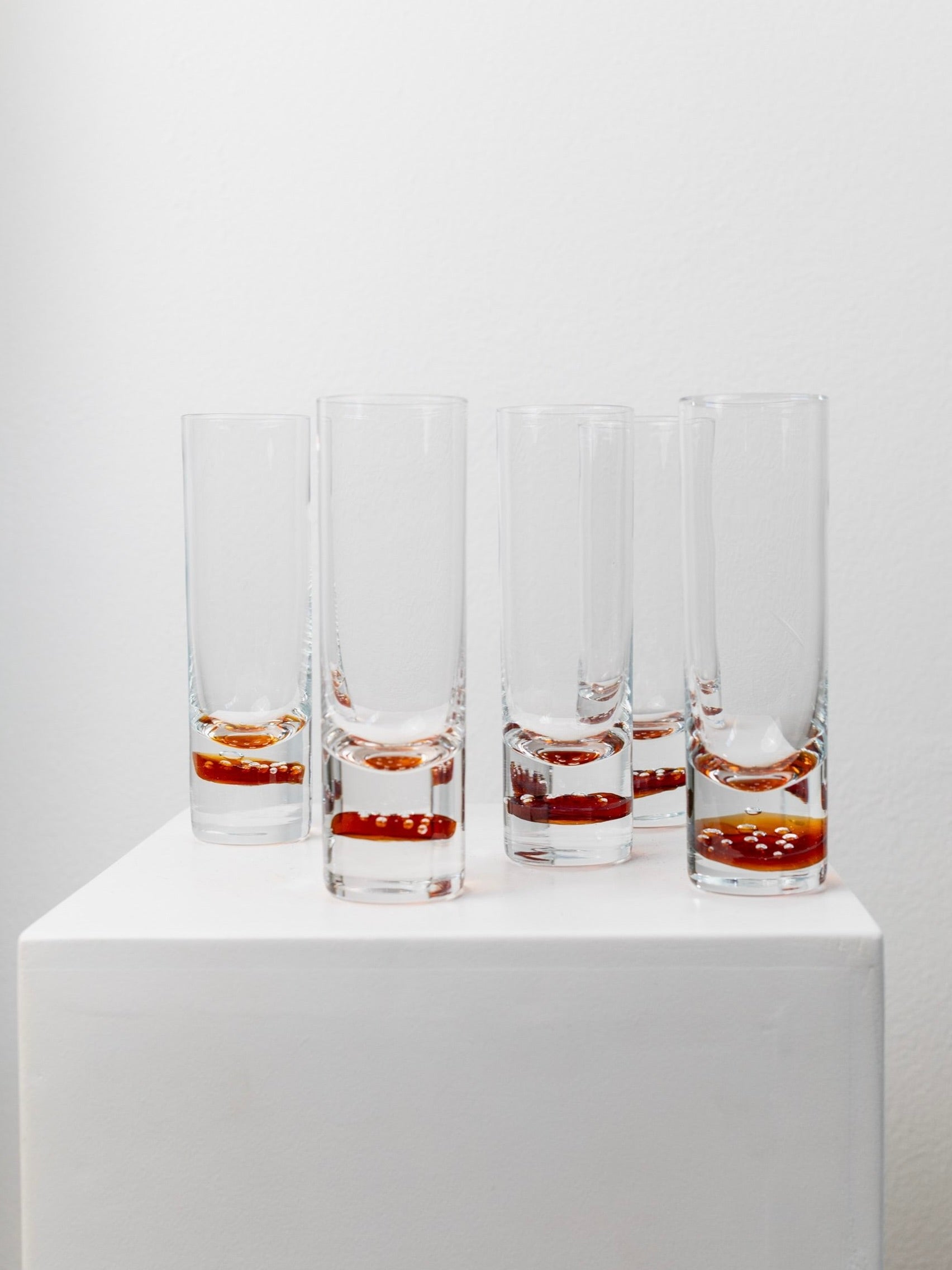 Arranged on a white pedestal against a plain white background are five tall, clear glasses from the Bottega Jacobs collection, each partially filled with a dark liquid. These 5 Retro Murano Drinking Glasses exude vintage charm with their mid-century design.