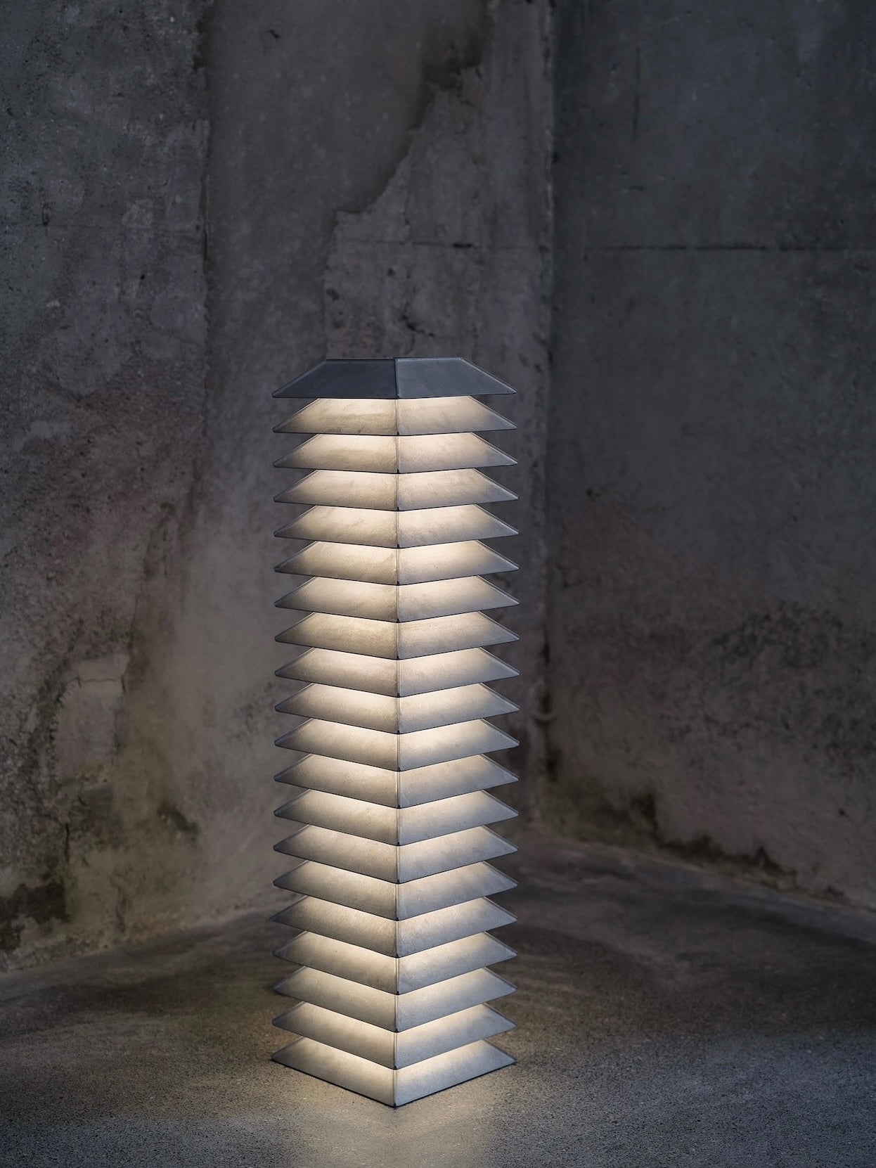 The Pagoda Floor Lamp by FEBRERO SHOP is a tall, modern piece crafted from industrial die-cast aluminum, emitting a soft white glow. It's part of a modular collection and stands in a dimly lit room with textured, unfinished walls, showcasing its geometric structure.