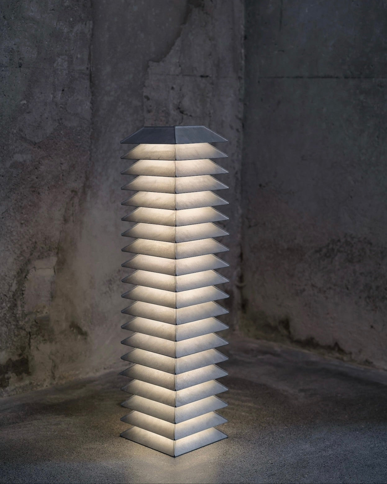 The Pagoda Floor Lamp by FEBRERO SHOP is a tall, modern piece crafted from industrial die-cast aluminum, emitting a soft white glow. It's part of a modular collection and stands in a dimly lit room with textured, unfinished walls, showcasing its geometric structure.
